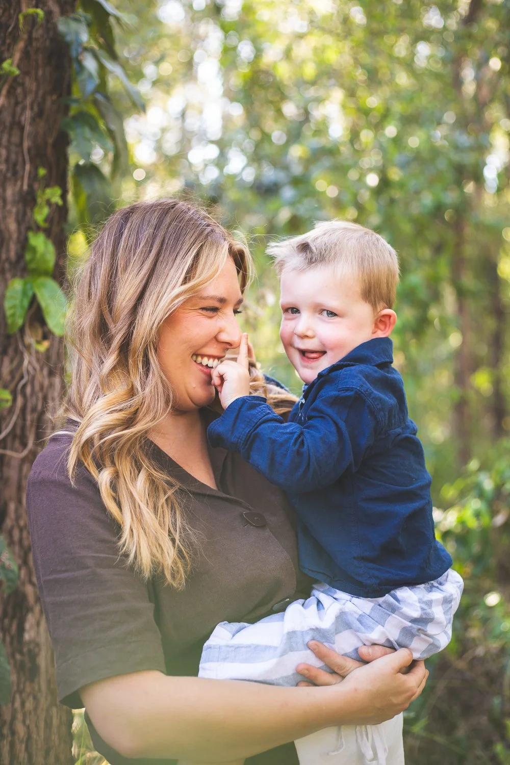 A smiling woman holding a young boy in a forested area, the boy touching her nose and both looking happy.