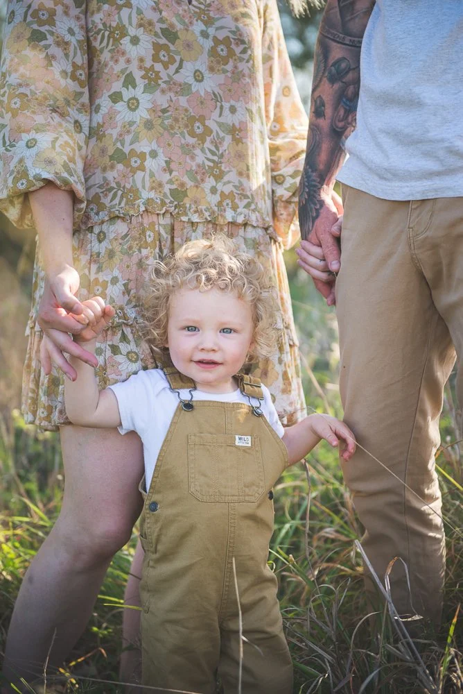 A young child with curly blonde hair and blue eyes standing outdoors, holding hands with two adults who are partially visible. The child is wearing tan overalls and a white shirt, with grass and trees in the background.