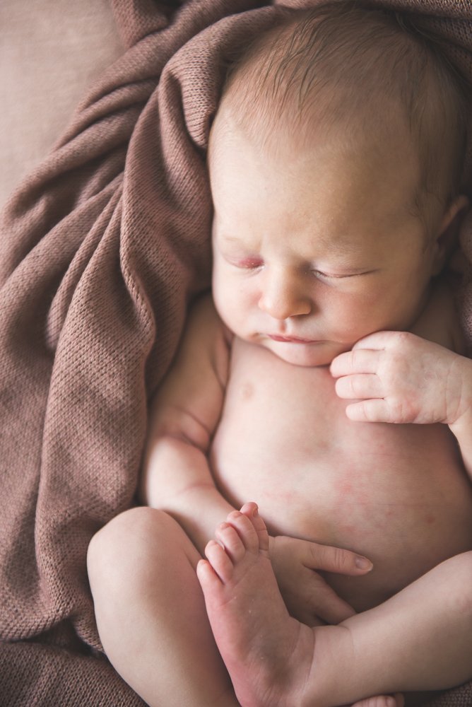 A newborn baby peacefully sleeping on a brown blanket, partially wrapped in a matching blanket, with a hand near the face.