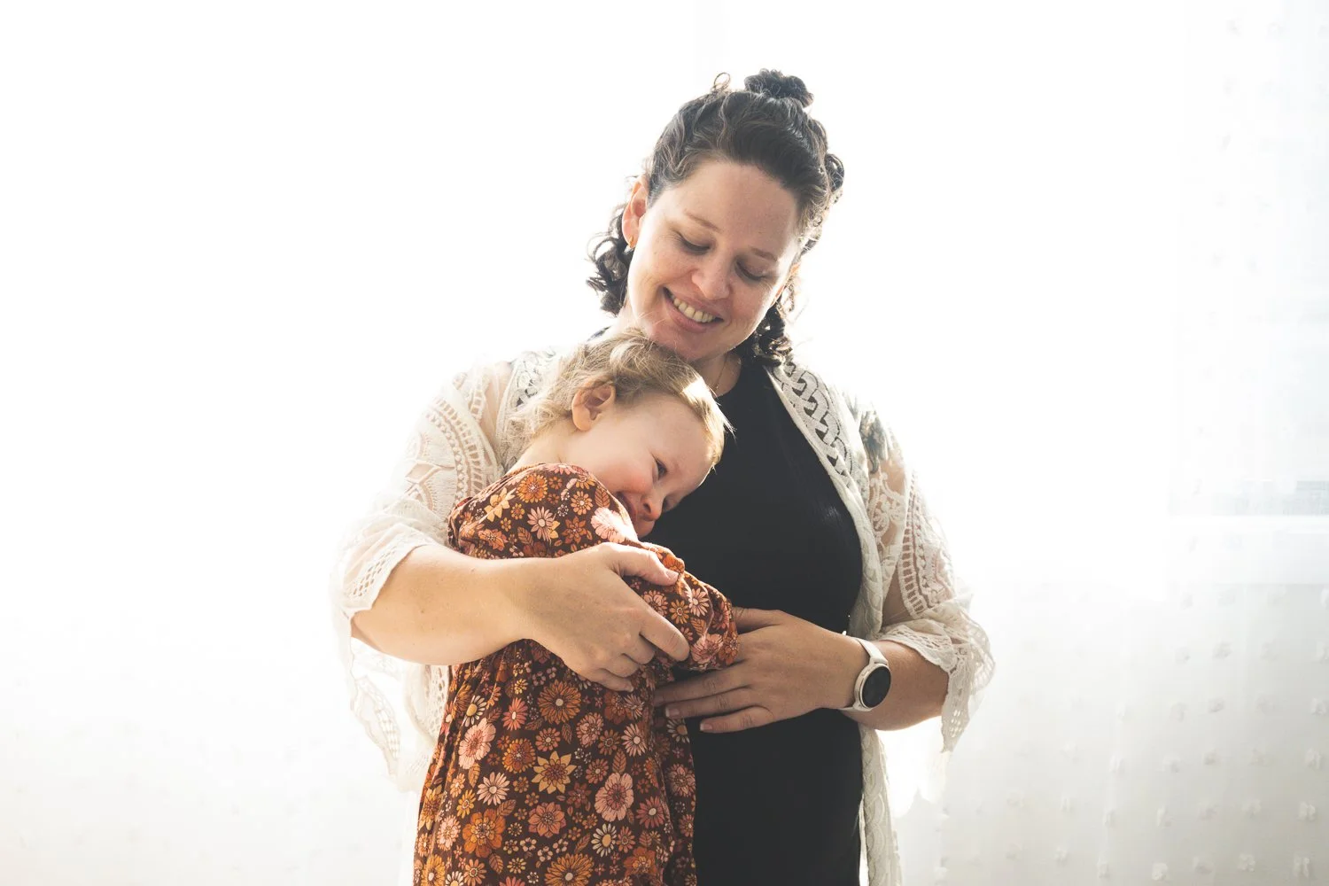 A woman and a young girl hugging and smiling, with the woman wearing a black dress and a white lace cardigan, and the girl wearing a floral dress, in front of a bright background.