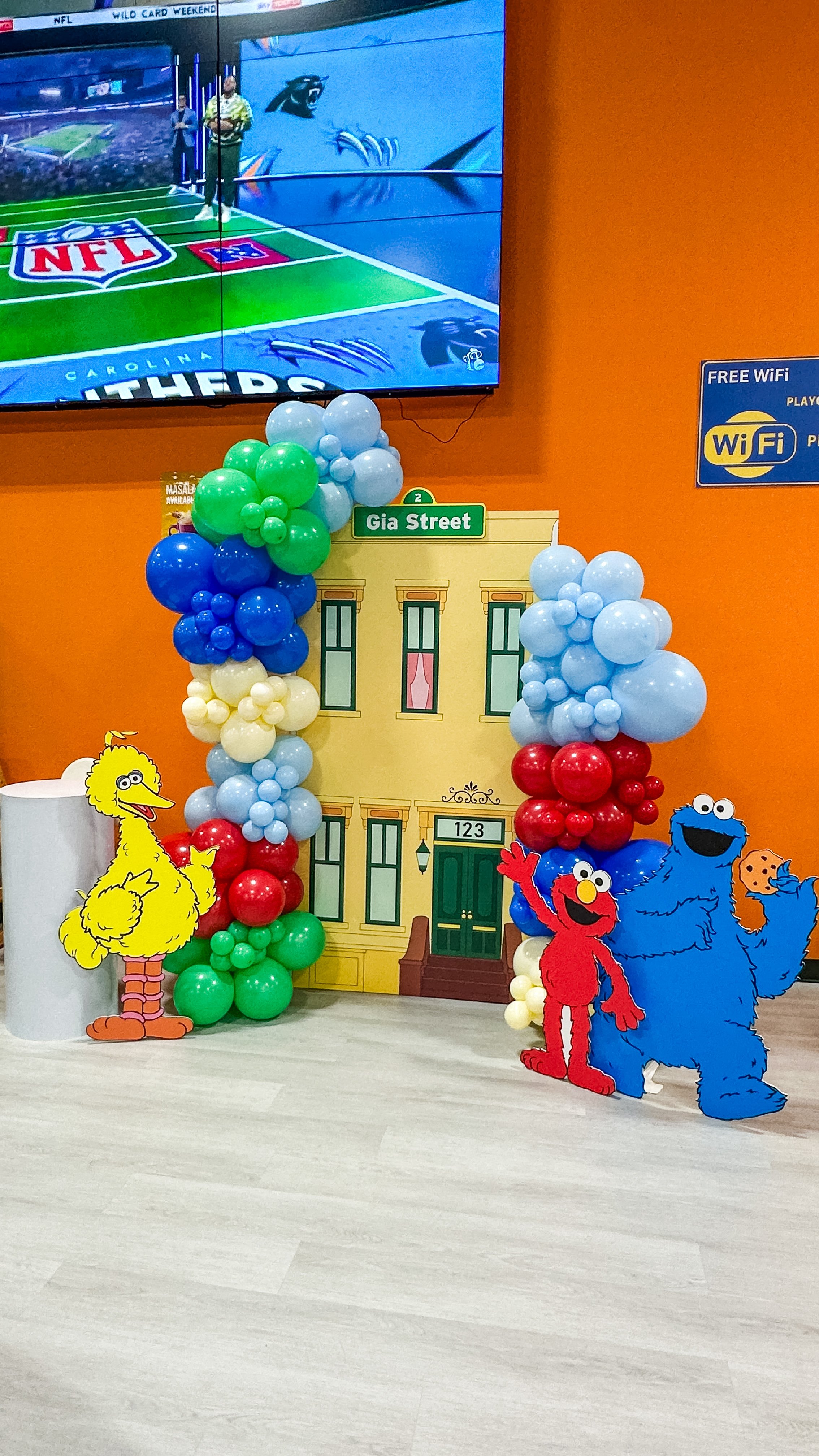 Decorative display of Sesame Street characters with balloons framing a background of a colorful apartment building and street sign that says 'Gia Street'. Animated characters Big Bird, Elmo, and Cookie Monster are cutouts positioned in front of the d