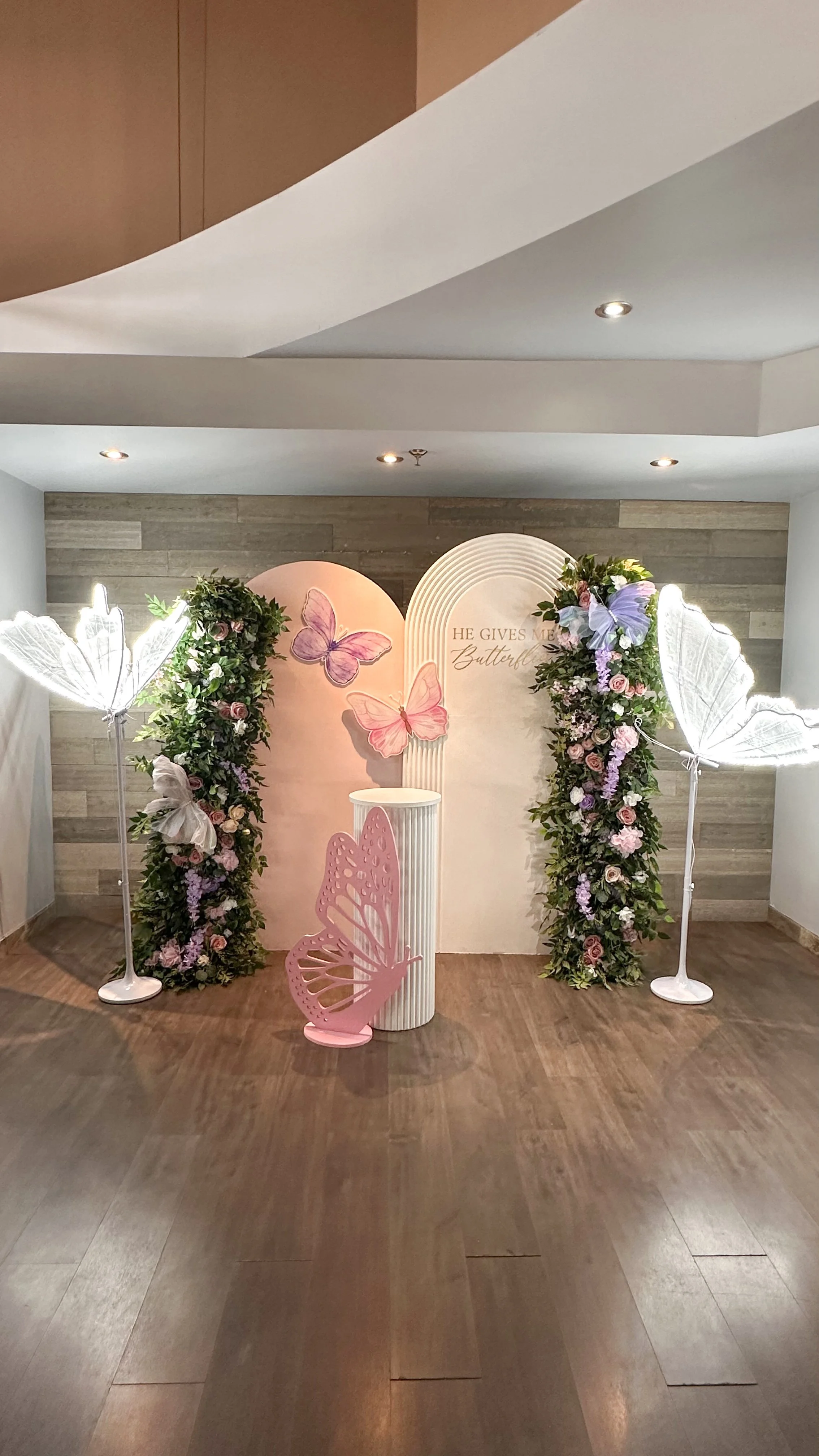 A decorative backdrop with pink butterflies and flowers, two white butterfly-shaped lights, and a sign that reads 'He gives me butterflies,' surrounded by floral arrangements.