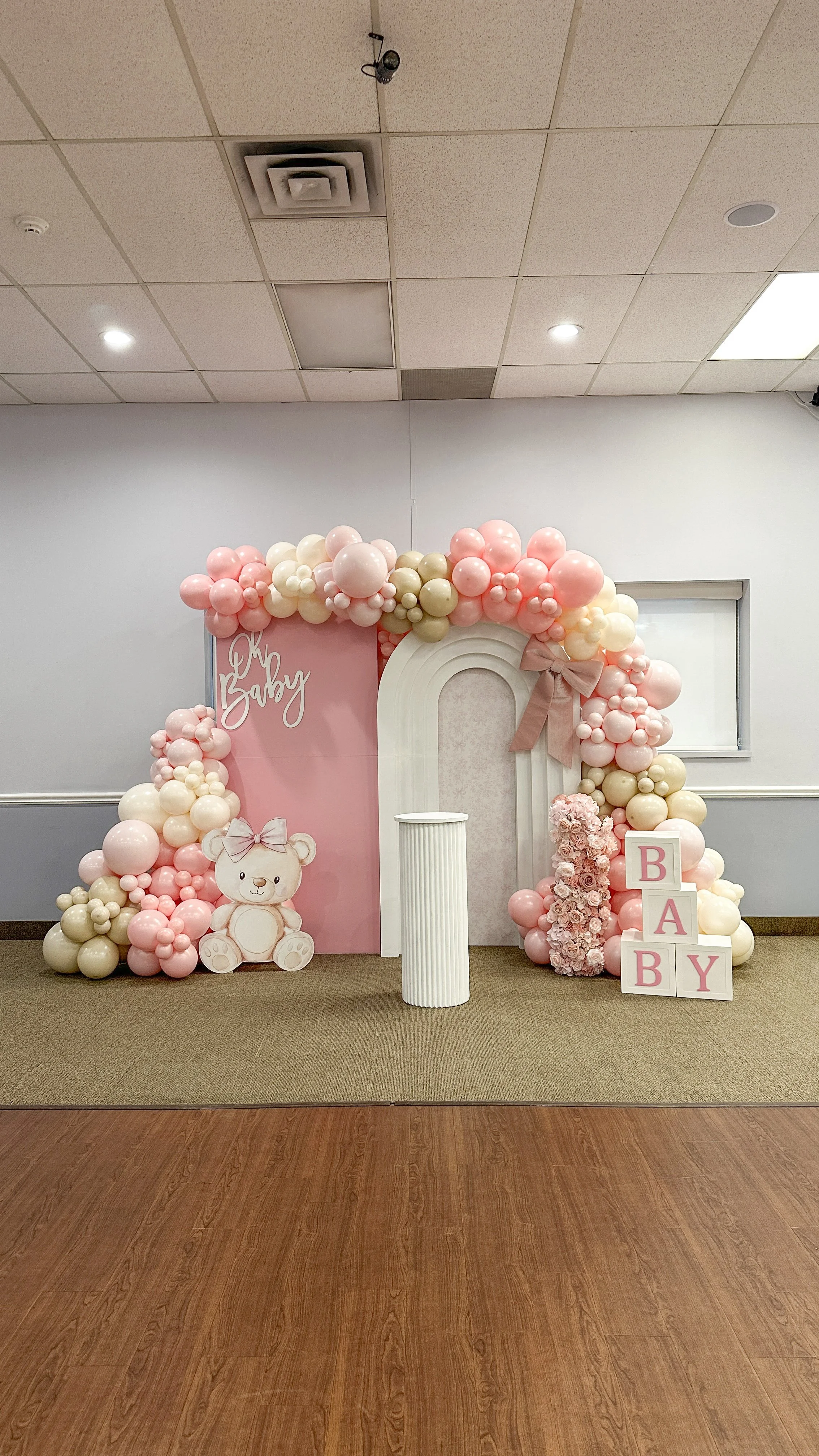 Baby shower decoration with pink, cream, and white balloons, a teddy bear cutout, a pink backdrop with the words 'Oh Baby,' and blocks spelling 'BABY'.