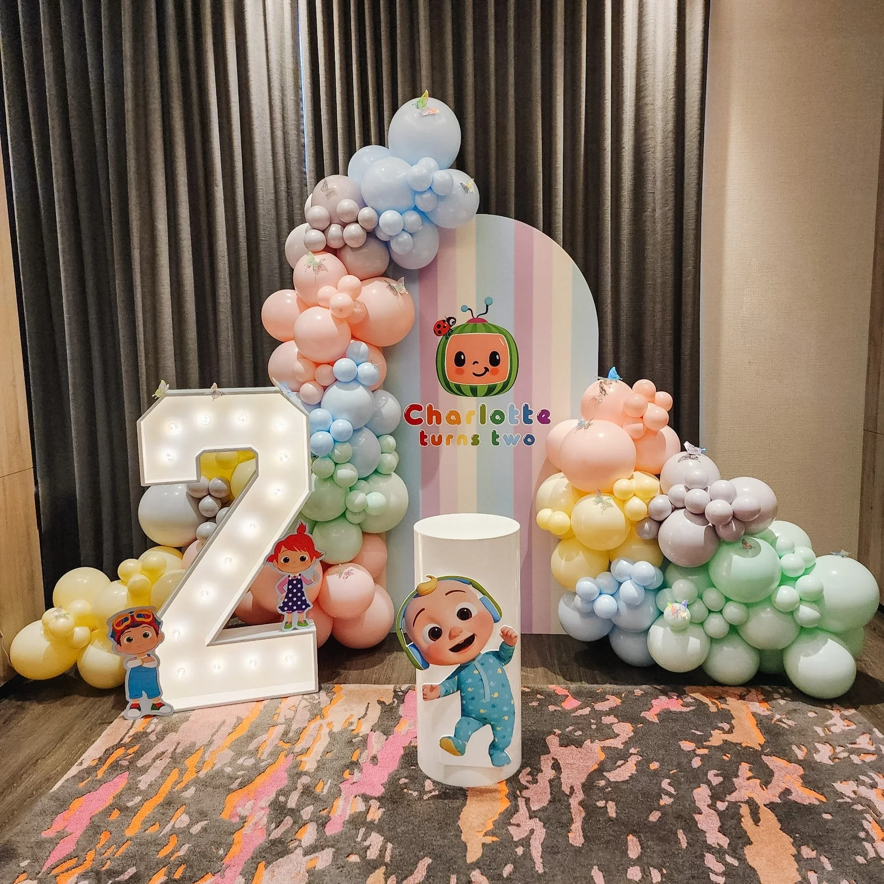 Colorful birthday party display for Charlotte's second birthday with pastel balloon arrangements, a large illuminated number 2, and cartoon character cutouts.