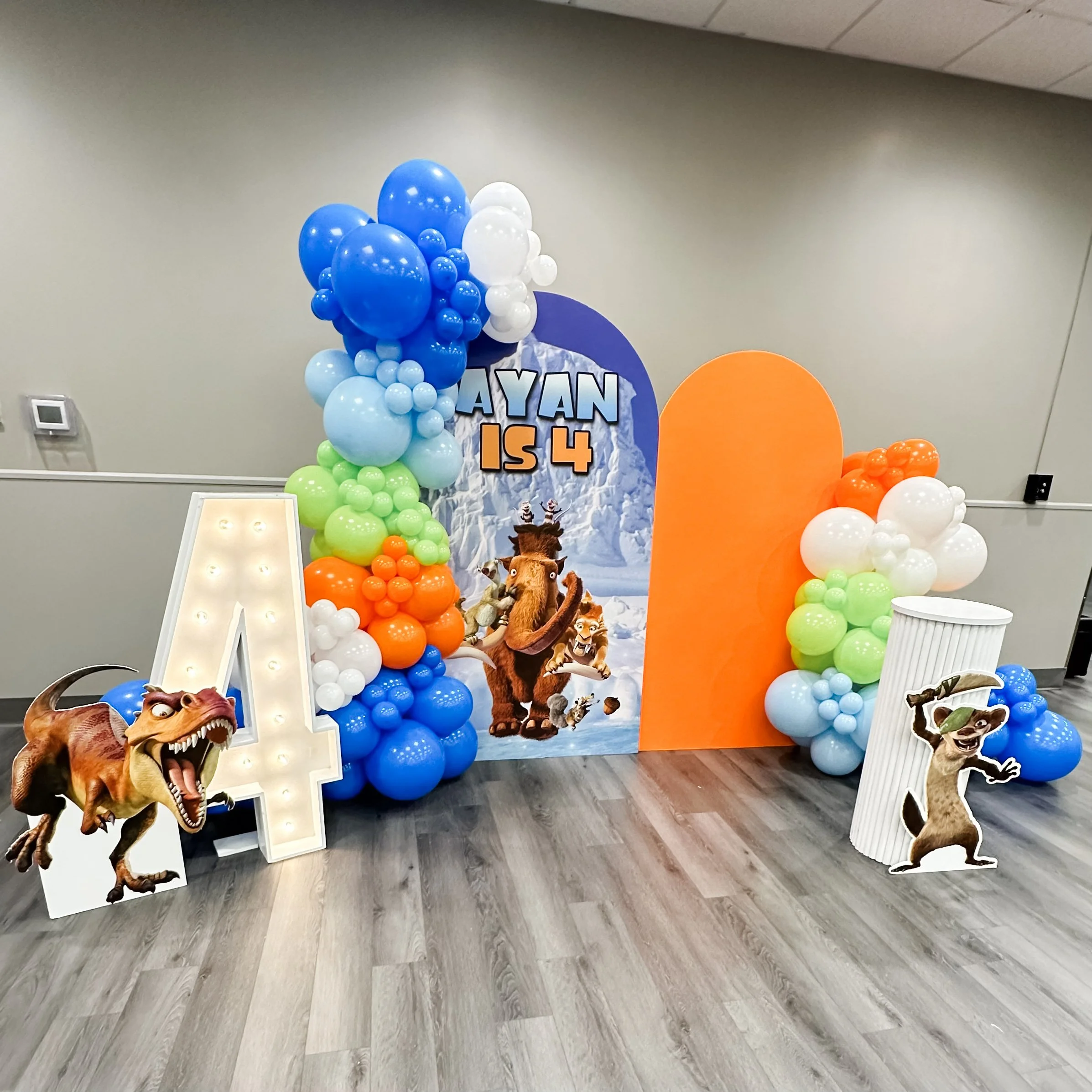Ice Age-themed birthday party backdrop with colorful balloons, large lit letter 'A', and characters from Ice Age. The backdrop displays the text 'AYAN IS 4' and features animals like a dinosaur, a squirrel, a sloth, and a saber-tooth tiger.