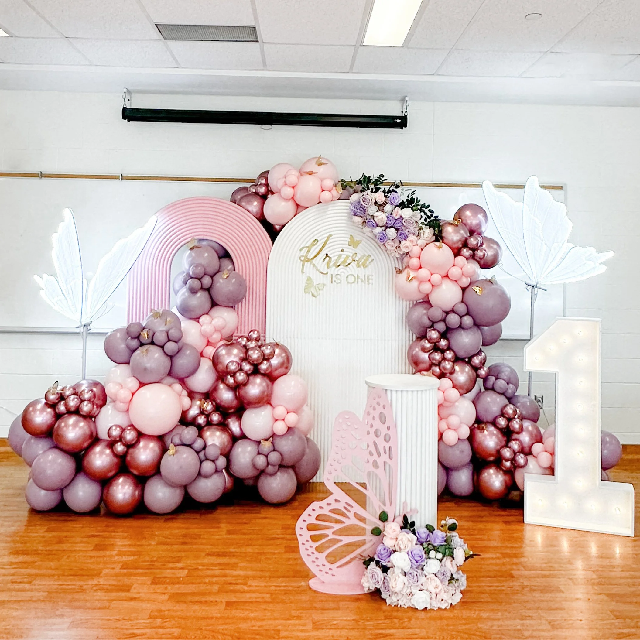 Pink and purple balloon arrangement with white butterfly decorations, a floral display, and a large illuminated number 1, set up for a first birthday celebration.