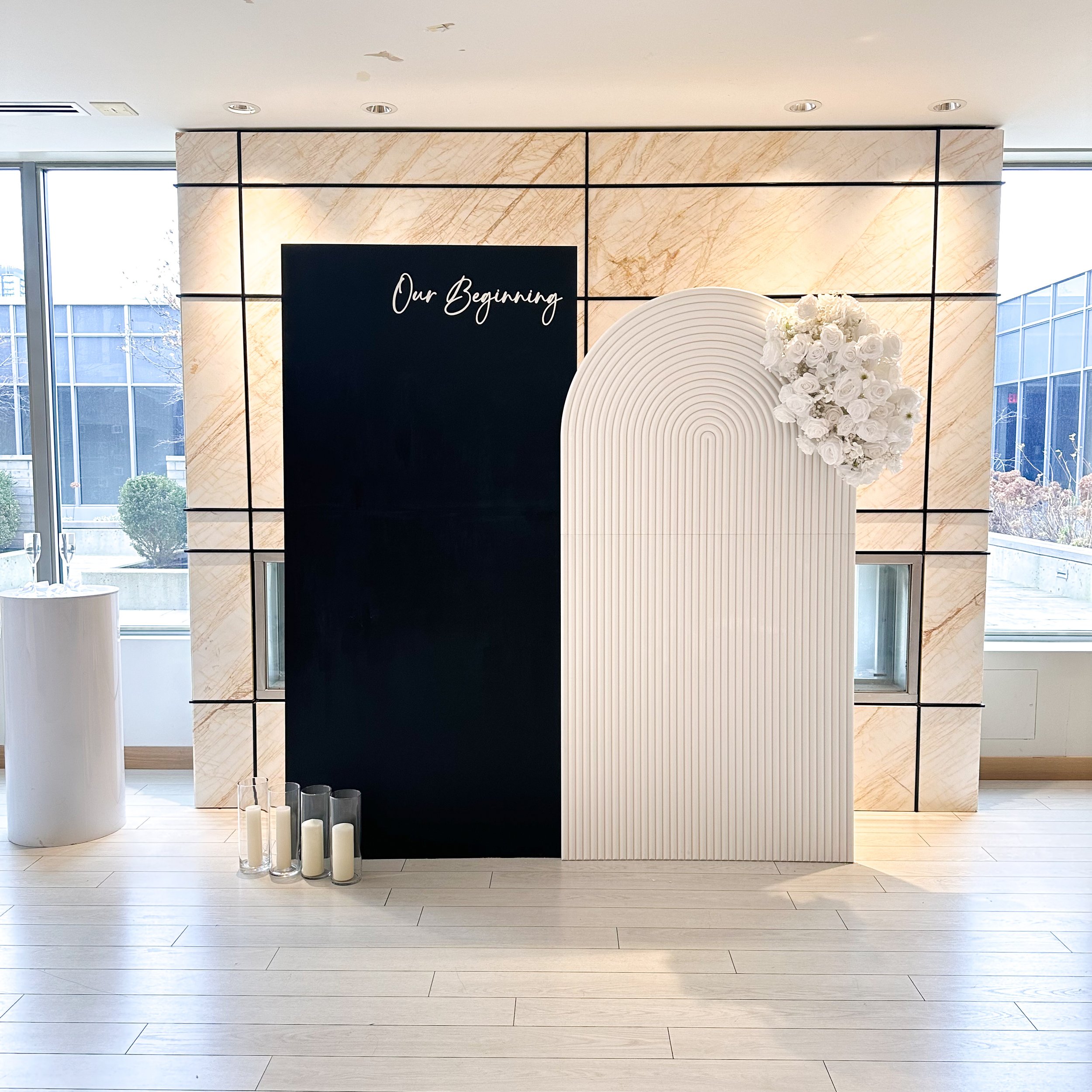 Minimalist wedding or event backdrop with black and white panels, a floral arrangement, and candles, with the text 'Our Beginning' on a black panel.