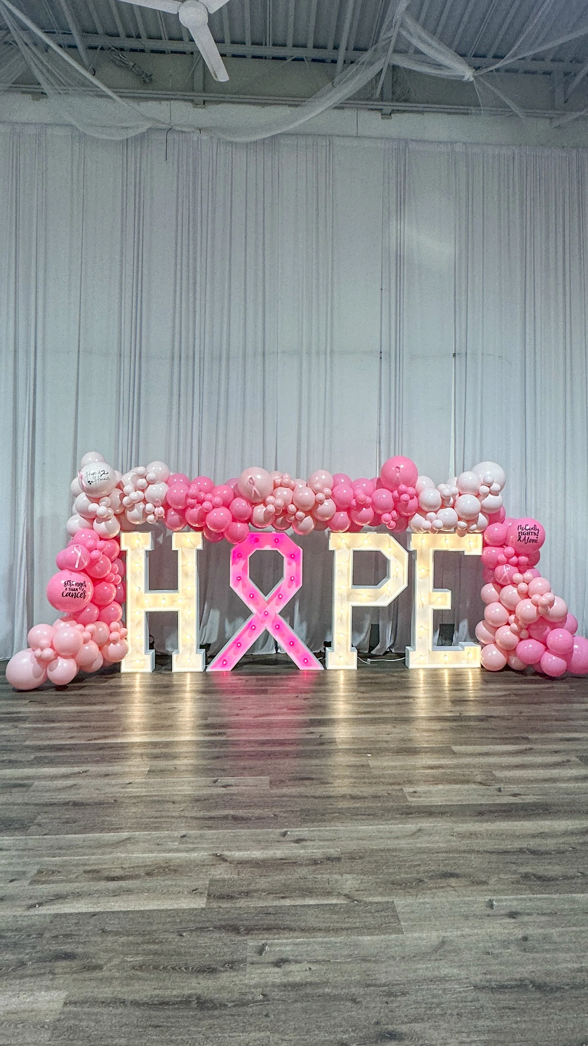 Decorative display with large illuminated letters spelling 'HOPE', surrounded by pink and white balloons, with a pink awareness ribbon in the center, on a wooden floor against a white curtain backdrop.
