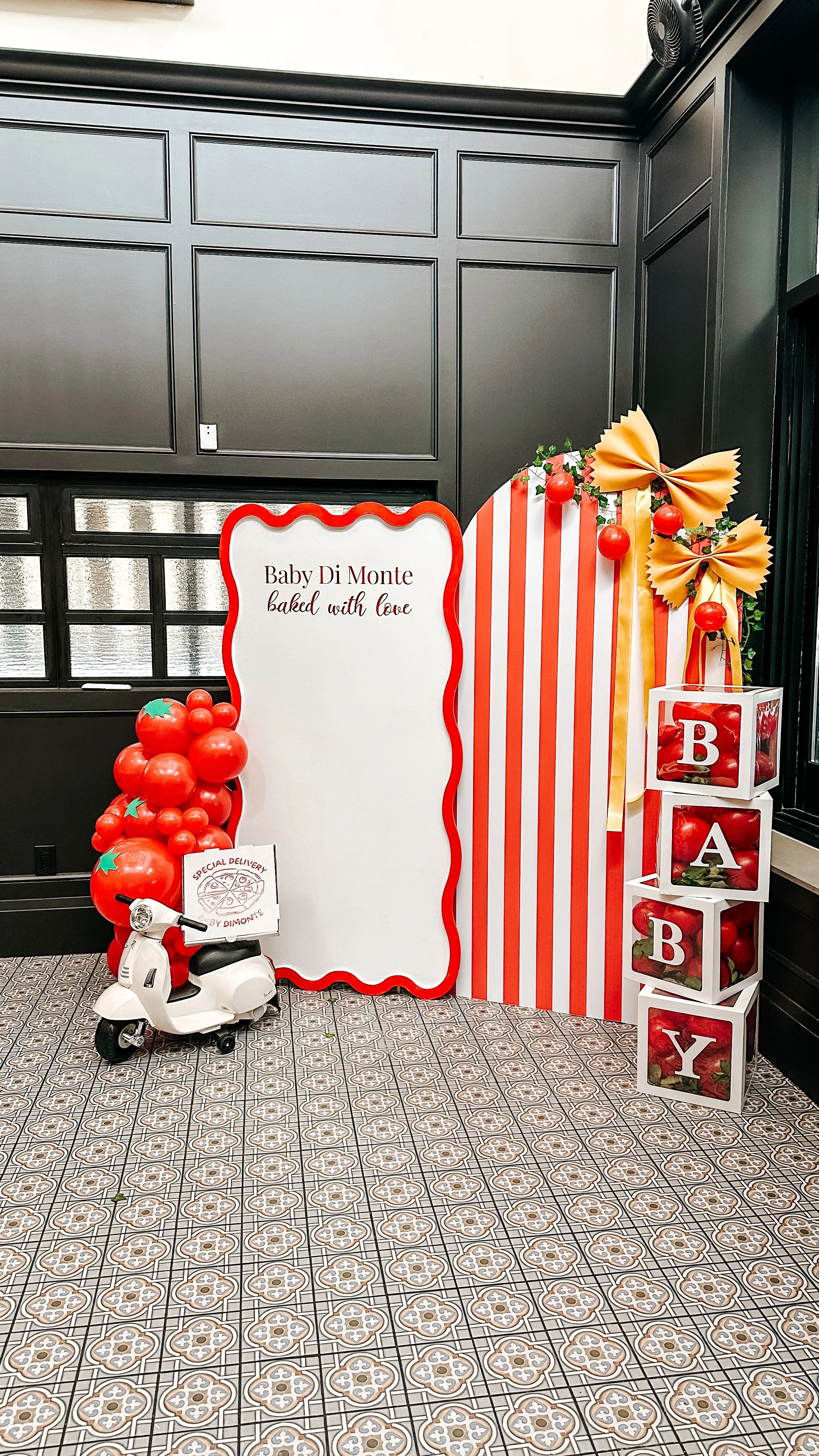 Decorative display with a white sign that reads 'Baby Di Monte baked with love', surrounded by red balloons, a small white scooter, and a stack of four white cubes spelling 'BABY' with red and green accents, all against black paneled walls and patter
