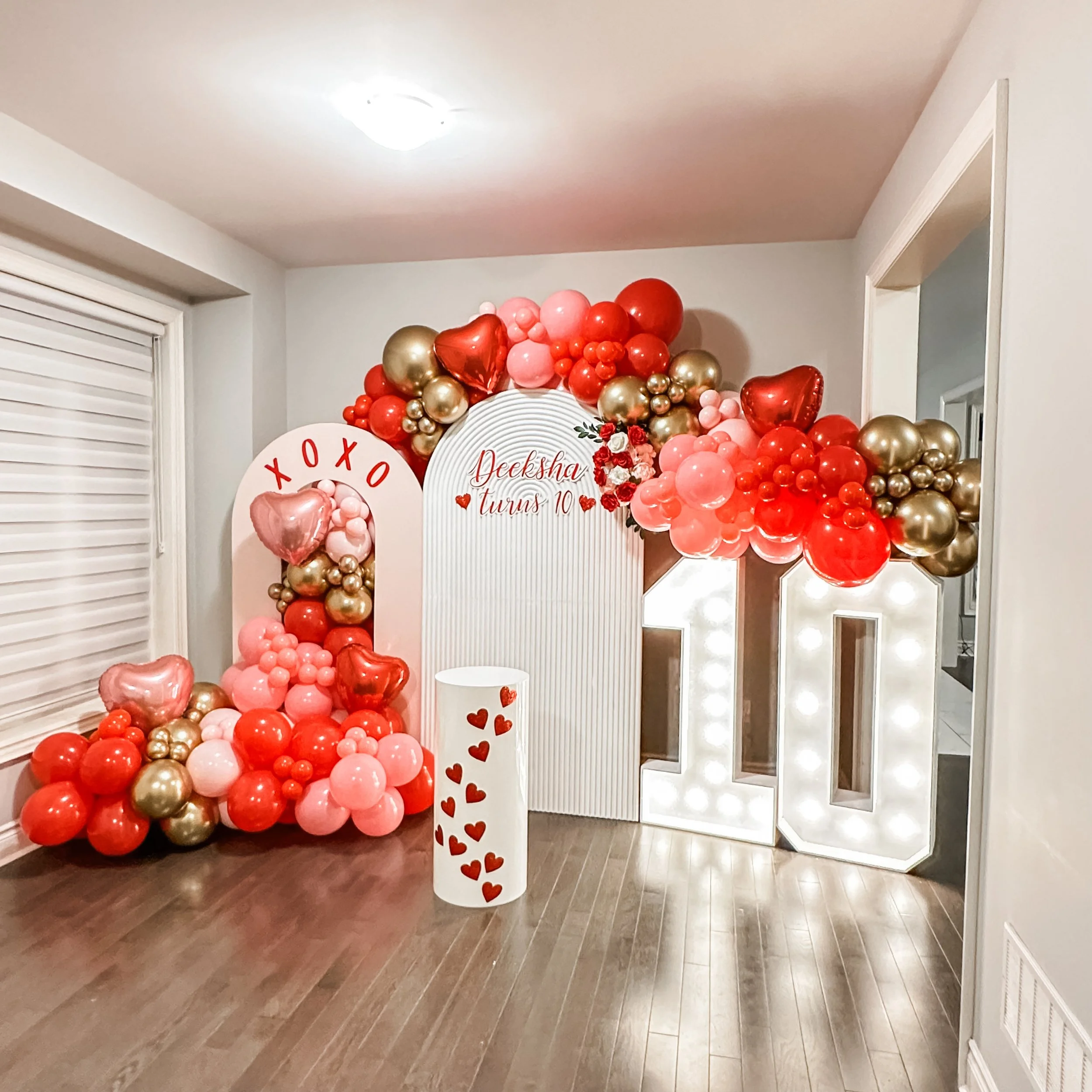 Valentine's Day themed balloon arrangement with red, pink, gold balloons, a large illuminated '10', and a white backdrop with red lettering that reads 'Decksha turns 10'.