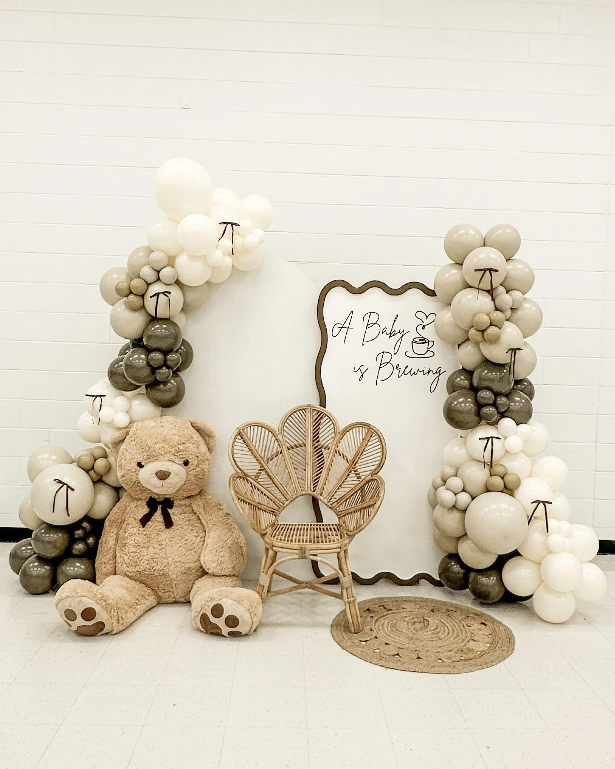 Balloon arch with white, beige, and gray balloons surrounds a white backdrop with a sign reading "A Baby is Brewing," accompanied by a wicker chair, a large teddy bear, and a round woven rug.