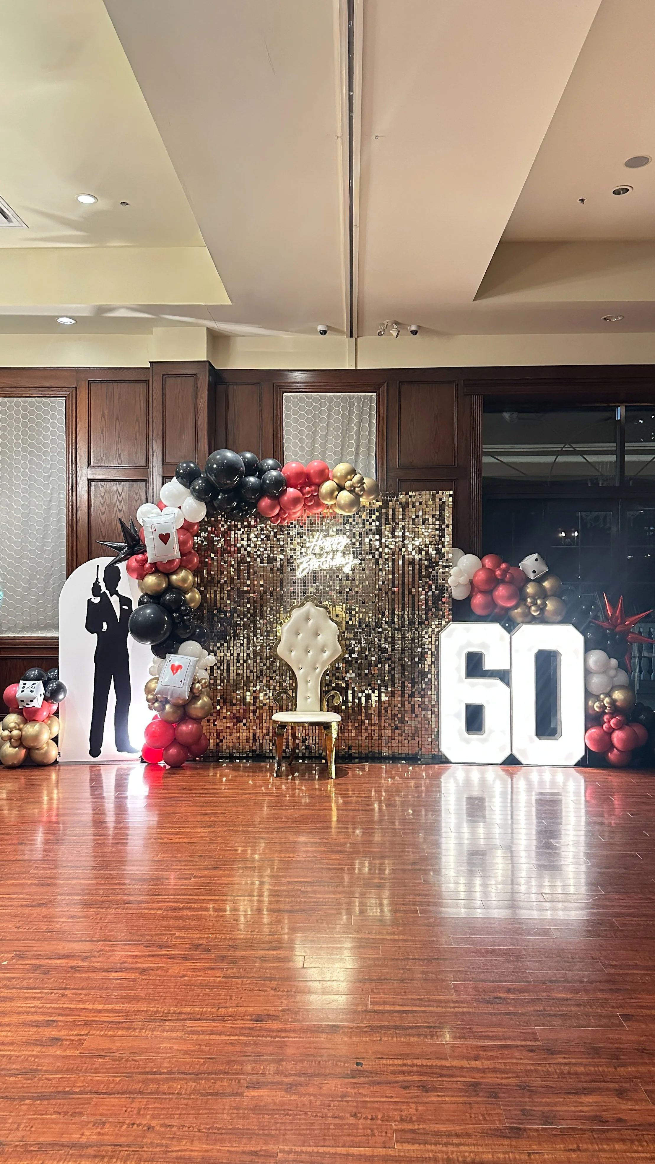 Party decoration setup with a golden twenty-five sign, a white ornate chair, and themed balloon arch and arrangements in black, gold, red, white, with a sequin backdrop and a silhouette of a man in a suit.