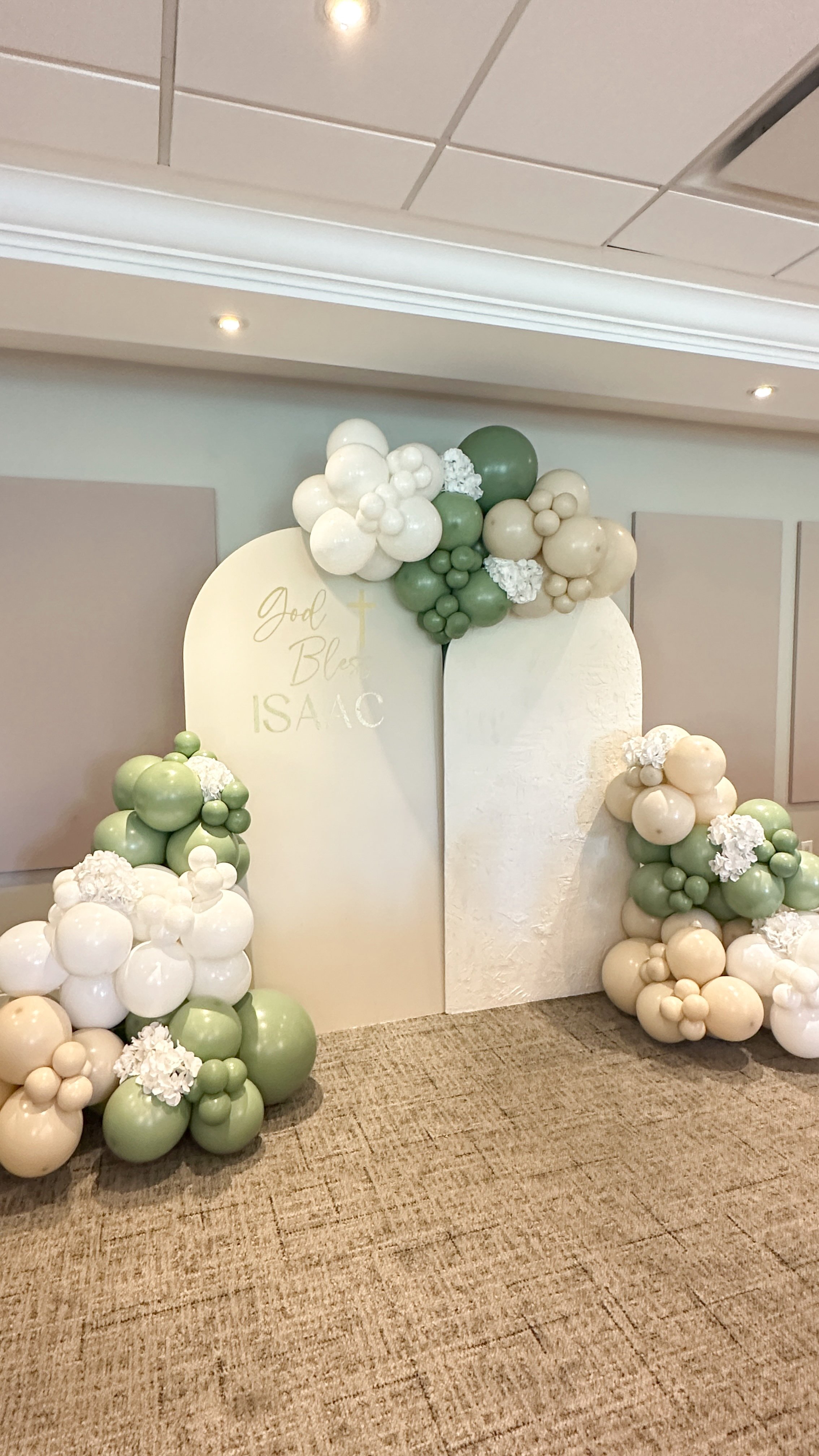 Decorative backdrop with white and green balloons, flowers, and a white sign that reads 'God Bless ISAAC'.