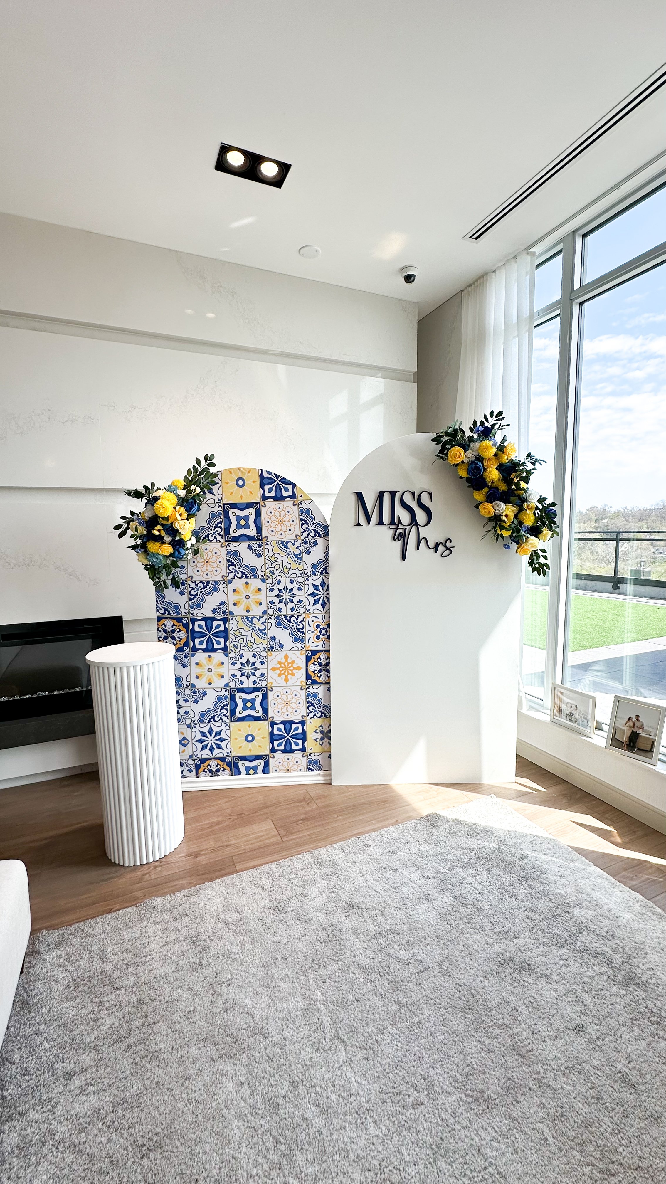 Decorative photo backdrop with yellow and blue floral arrangements and a tile pattern, labeled 'Miss to Mrs', set in a bright room with large windows.