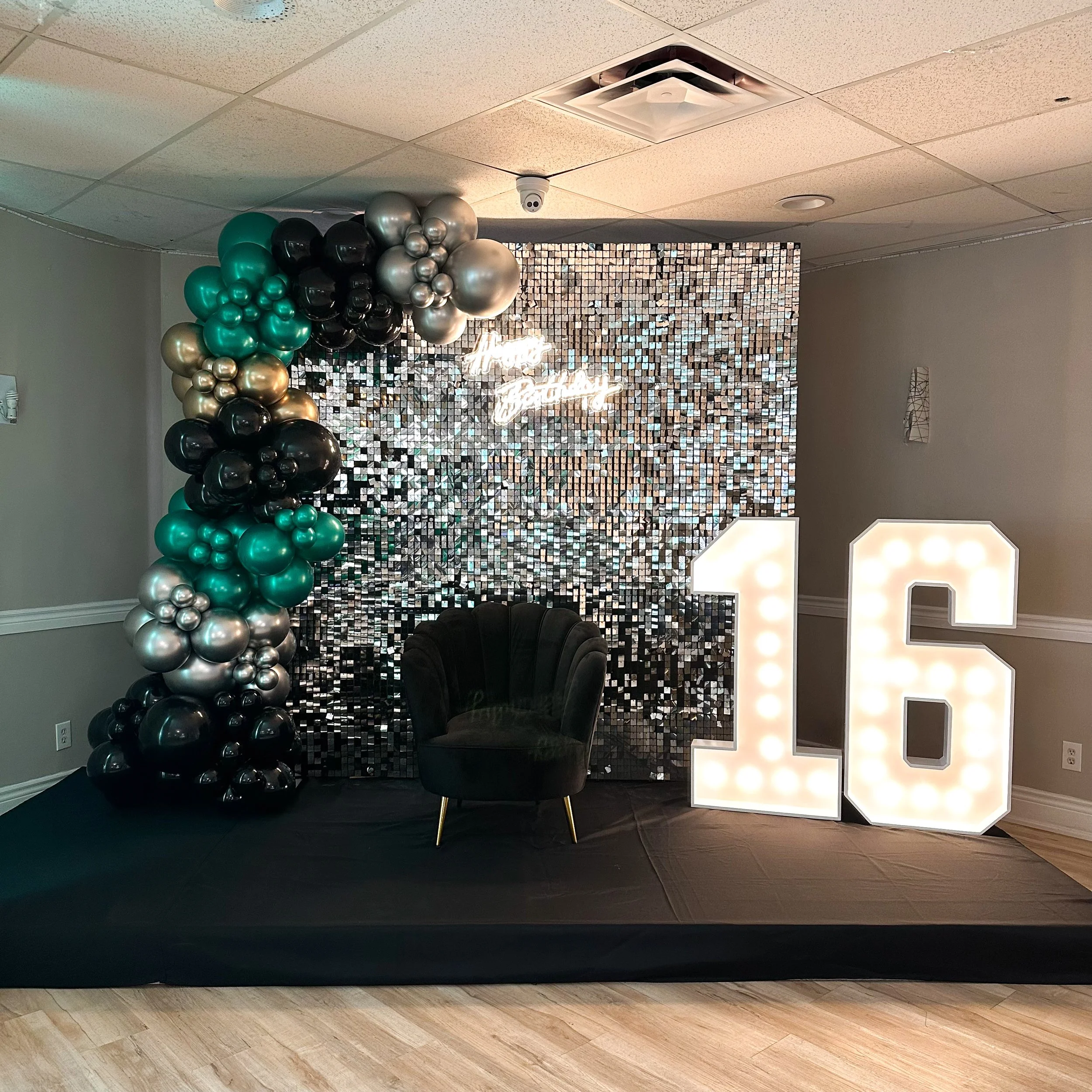 Decorative setup for a 16th birthday celebration featuring a large illuminated number 16, a black velvet chair, a backdrop made of small mirror tiles, a balloon arrangement in black, teal, and silver, and a neon sign that says "Happy Birthday."