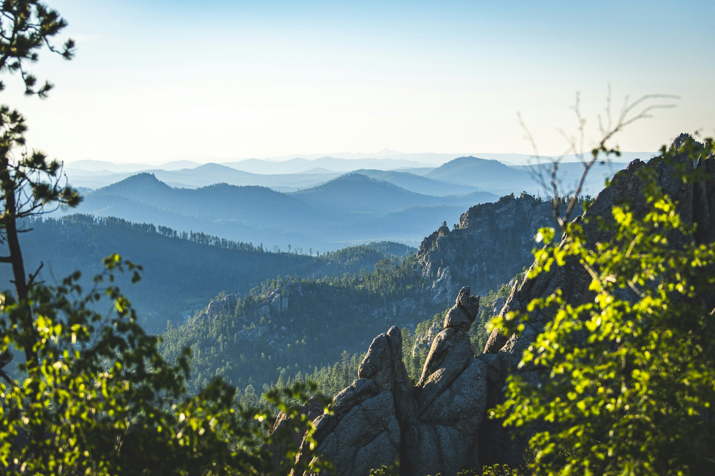 A scenic mountain landscape with layered blue mountains, rocky cliffs, and green trees, under a clear sky.