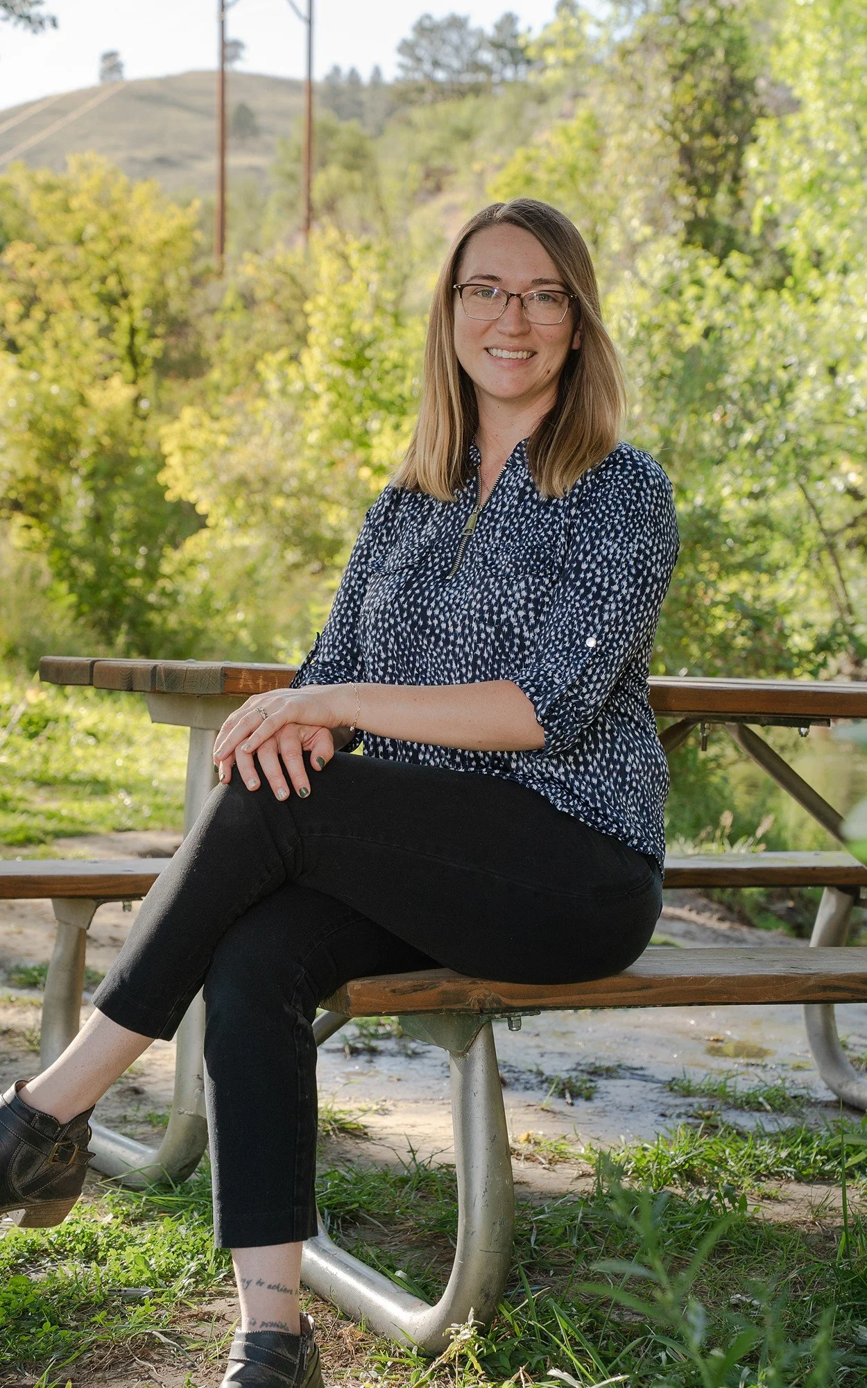 A young woman with glasses and long blonde hair sitting on a wooden park bench surrounded by green trees and yellowish foliage, smiling at the camera.