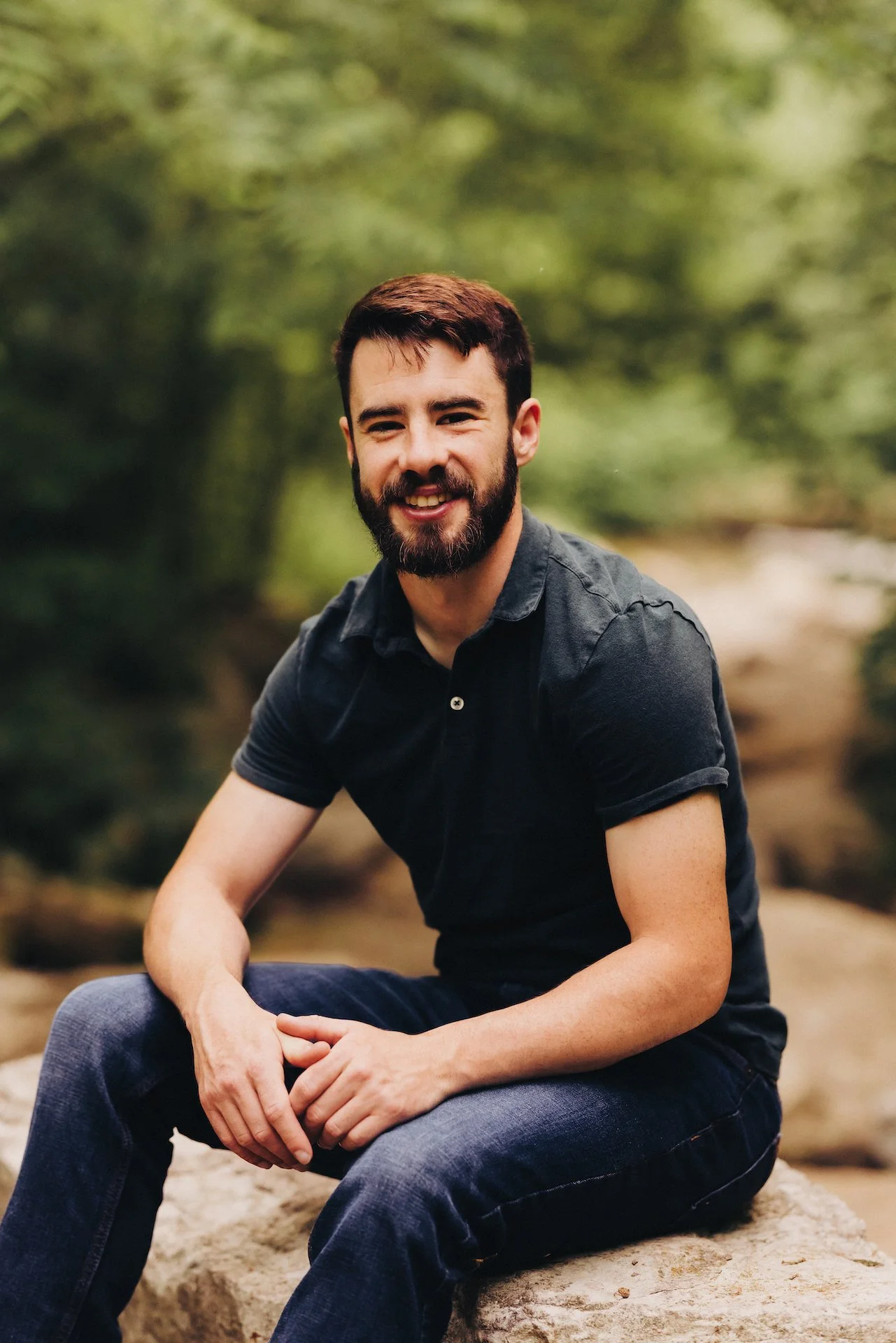 A young man with a beard, wearing a black polo shirt and jeans, sitting on a rock outdoors with a blurred green natural background.