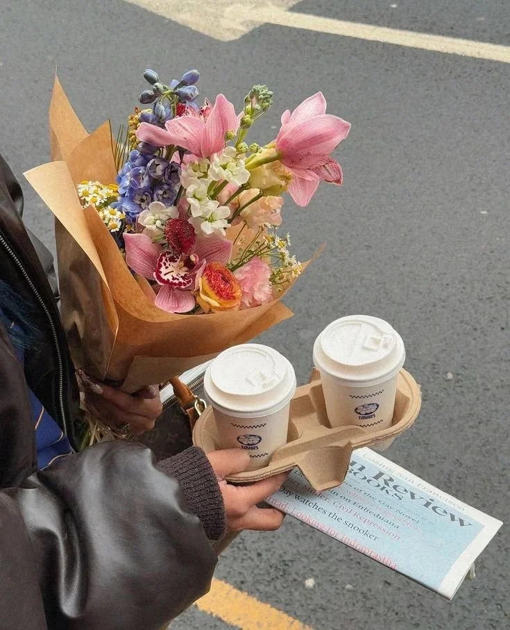 Person holding a bouquet of colorful flowers, two to-go coffee cups in a holder, and a newspaper, standing on a paved street.