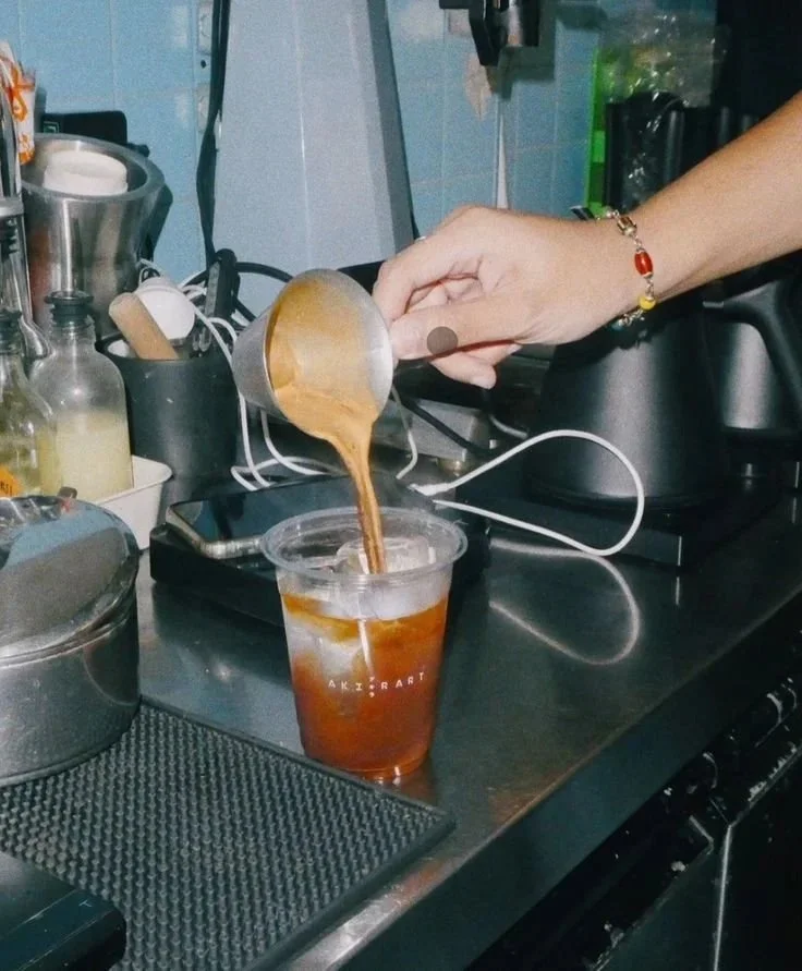 A person is pouring coffee from a metal pitcher into a plastic cup filled with ice and cold brew on a kitchen counter.