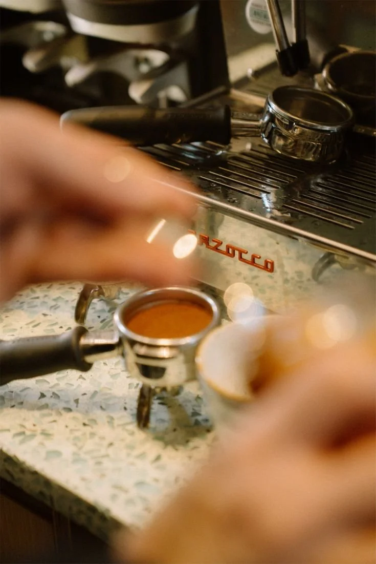 Close-up of a portafilter filled with espresso, held over a countertop in a coffee shop. A shiny espresso machine with the brand name 'Mazzerco' is in the background, with some equipment on top.
