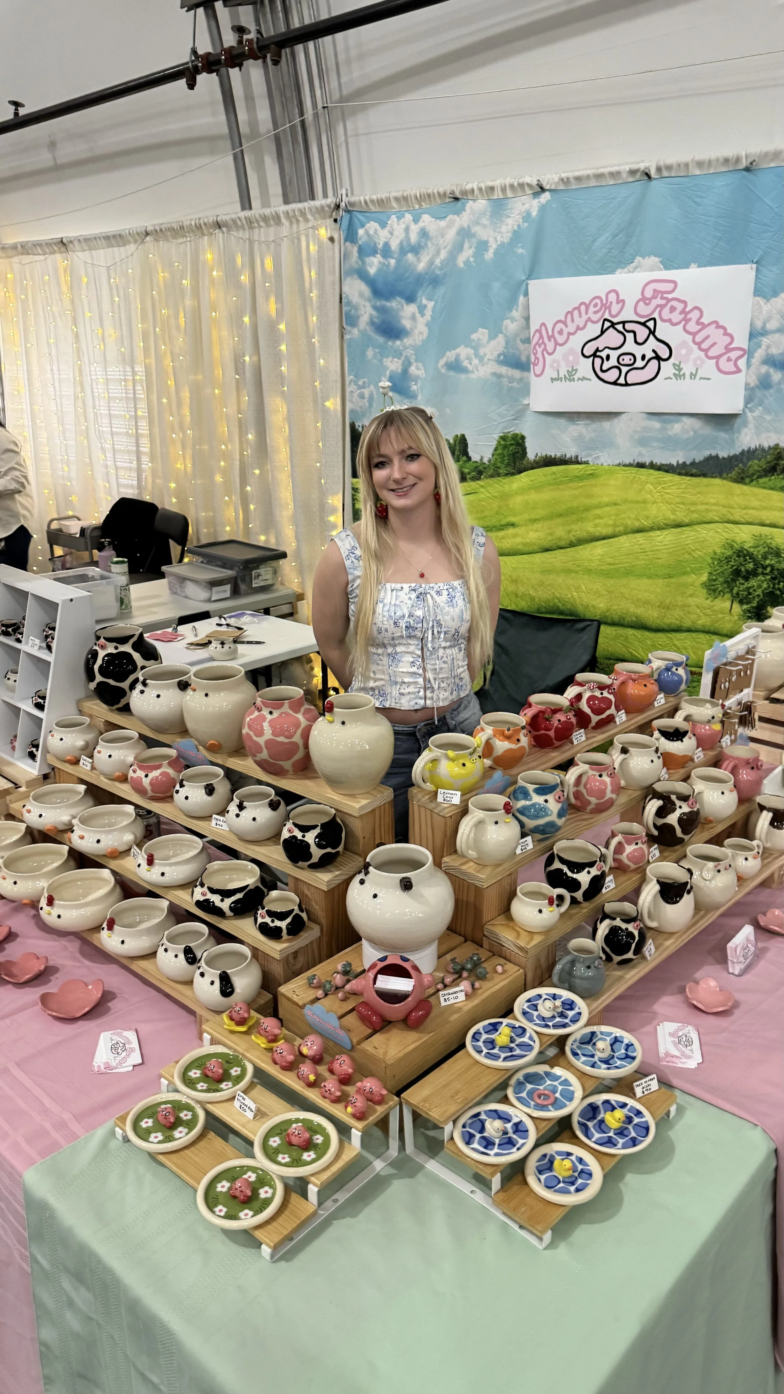 A woman standing behind a table of colorful ceramic pottery at a craft market, with a 'Flamer Farm' sign and a scenic backdrop in the background.