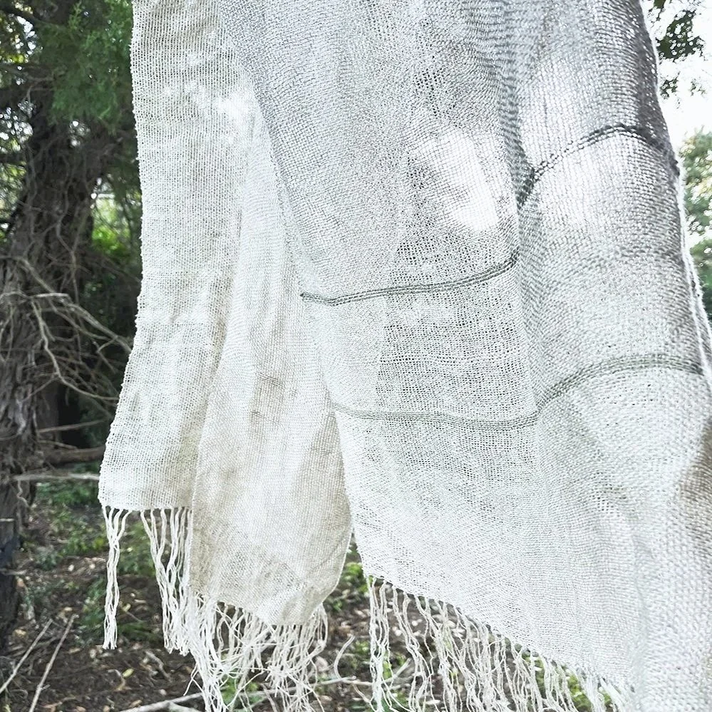 A close-up of a fine linen hand woven cloth with a fringe hanging on branch in the wind with trees and greenery visible in the background.