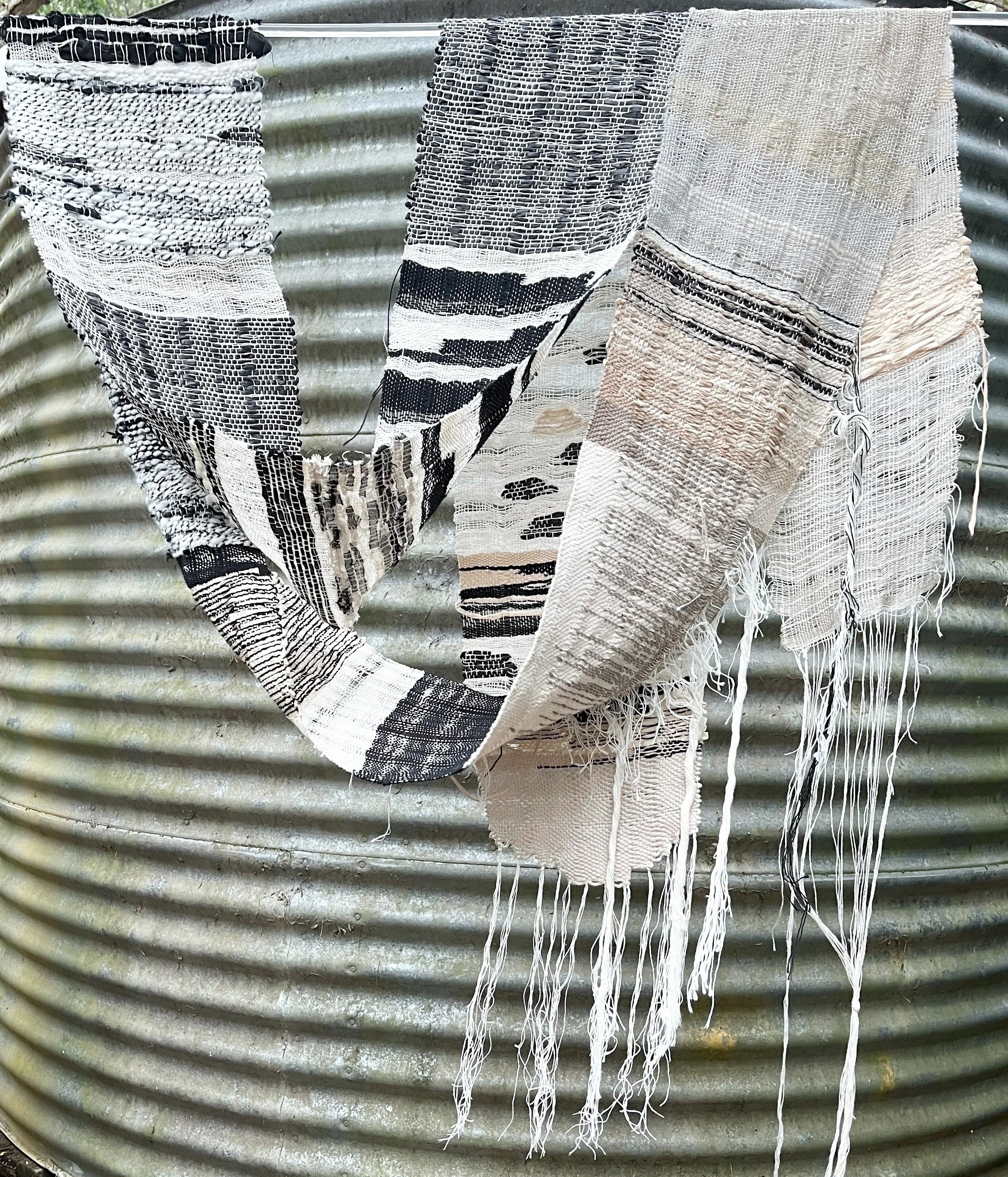 A long strip of handwoven textured textile made from paper, linen, hemp, wool and cotton hanging on a silver corrugated metal surface.