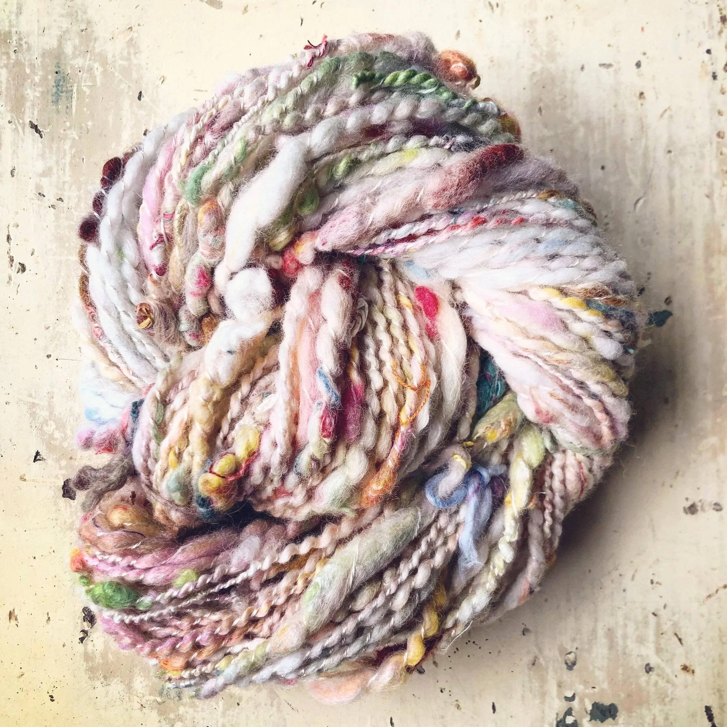 A ball of multicolored, textured yarn with shades of pink, white, green, yellow, and blue, resting on a worn, off-white surface.