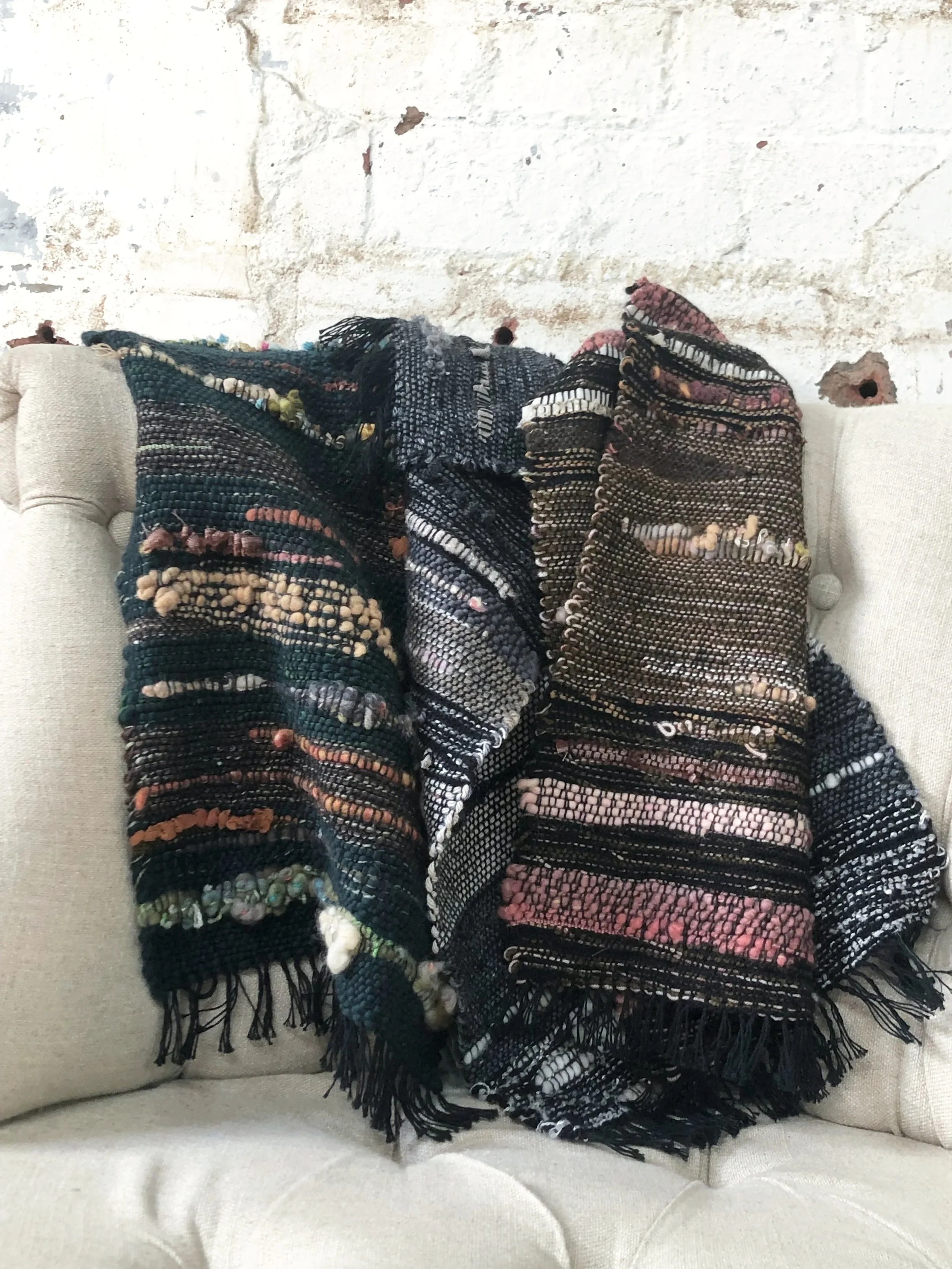 Colorful woven blanket draped over a beige couch against a textured white brick wall.