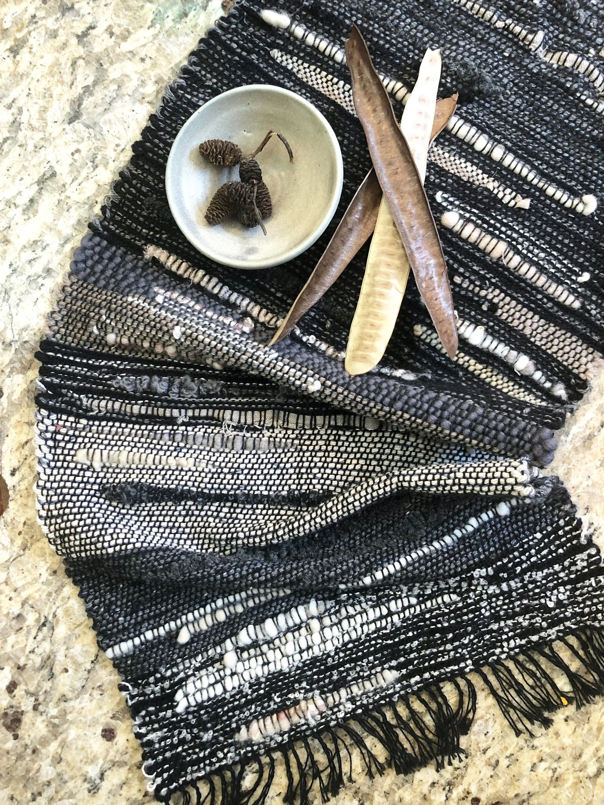A black and white woven textile with a small gray bowl containing three dried pine cones, and three dried seed pods or pods with a woody texture placed on the textile.