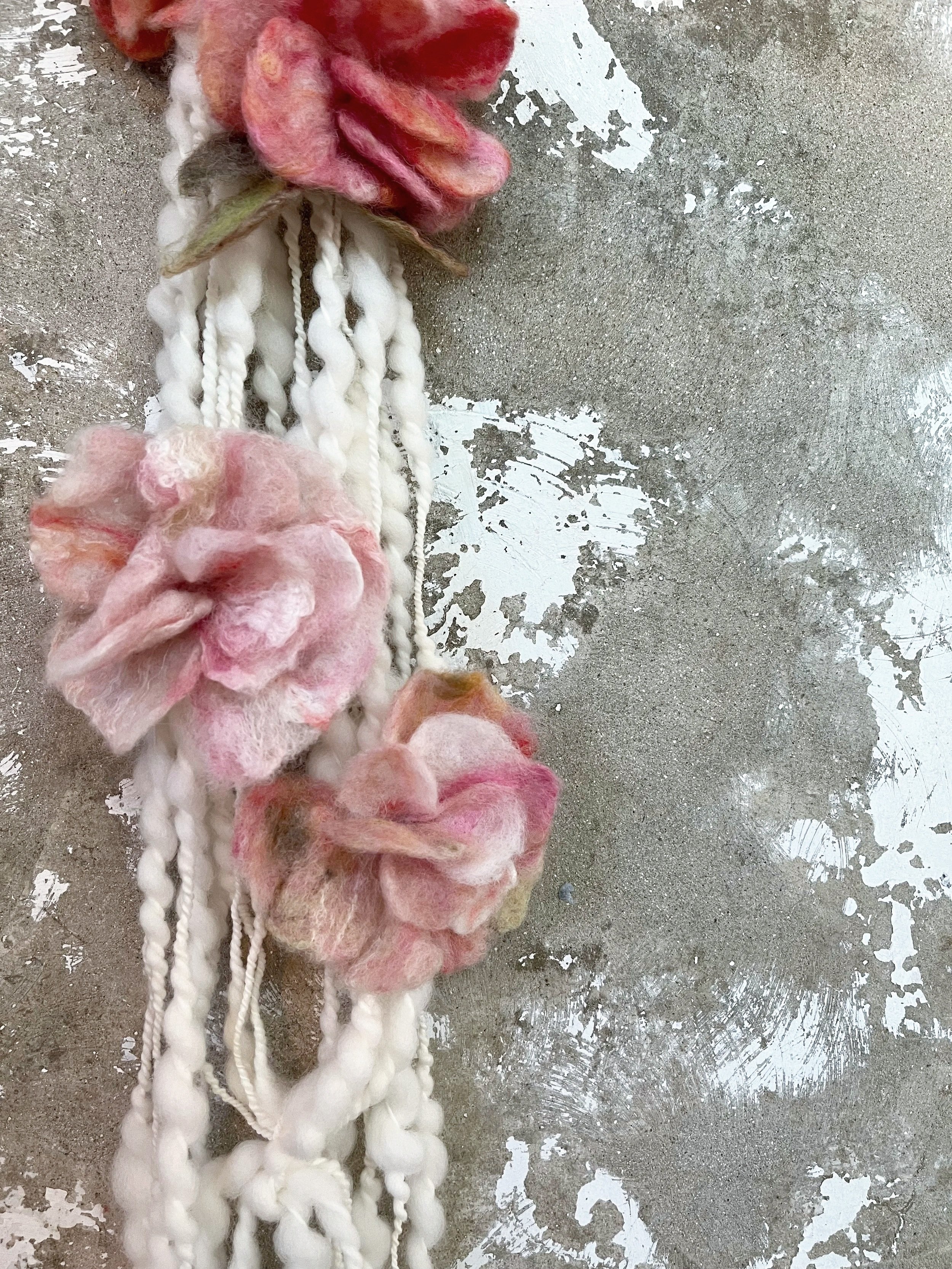 Handmade yarn necklace with felted pink flowers, laid on a distressed concrete surface.