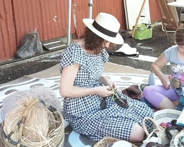 A woman with curly hair wearing a checkered dress and a wide-brimmed hat, sitting on a mat outdoors, holding art materials and surrounded by crafting supplies and woven baskets.