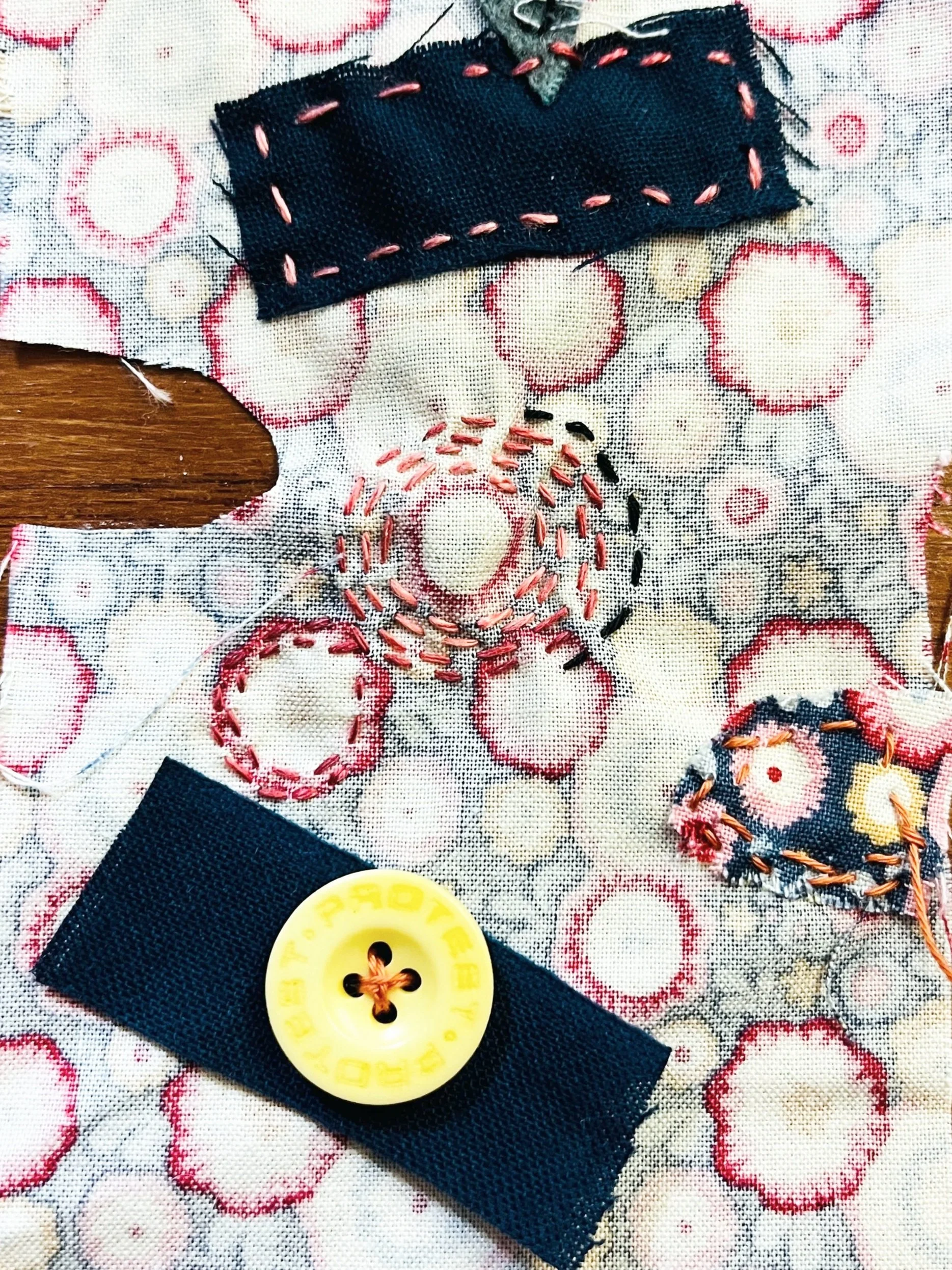 Close-up of fabric with stitched embroidery, showing black, pink, and multicolored threadwork, a yellow plastic button, and black fabric patches on a patterned cloth background.