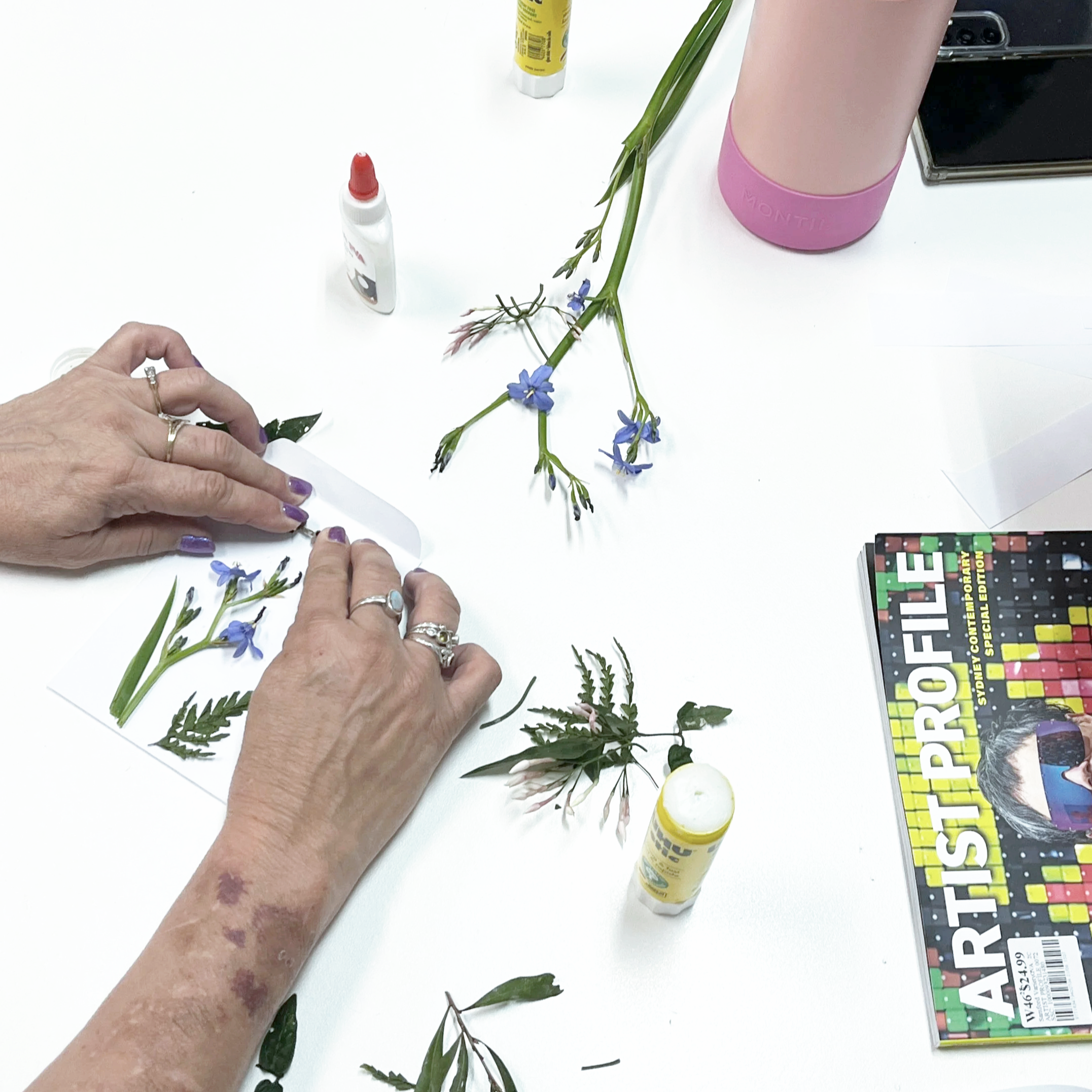 A person with purple nails is arranging small flowers and leaves on sheets of paper at a cluttered table. The table also has a pink water bottle, glue sticks, colorful pens, a magazine titled 'ARTIST PROFILE,' and a smartphone.