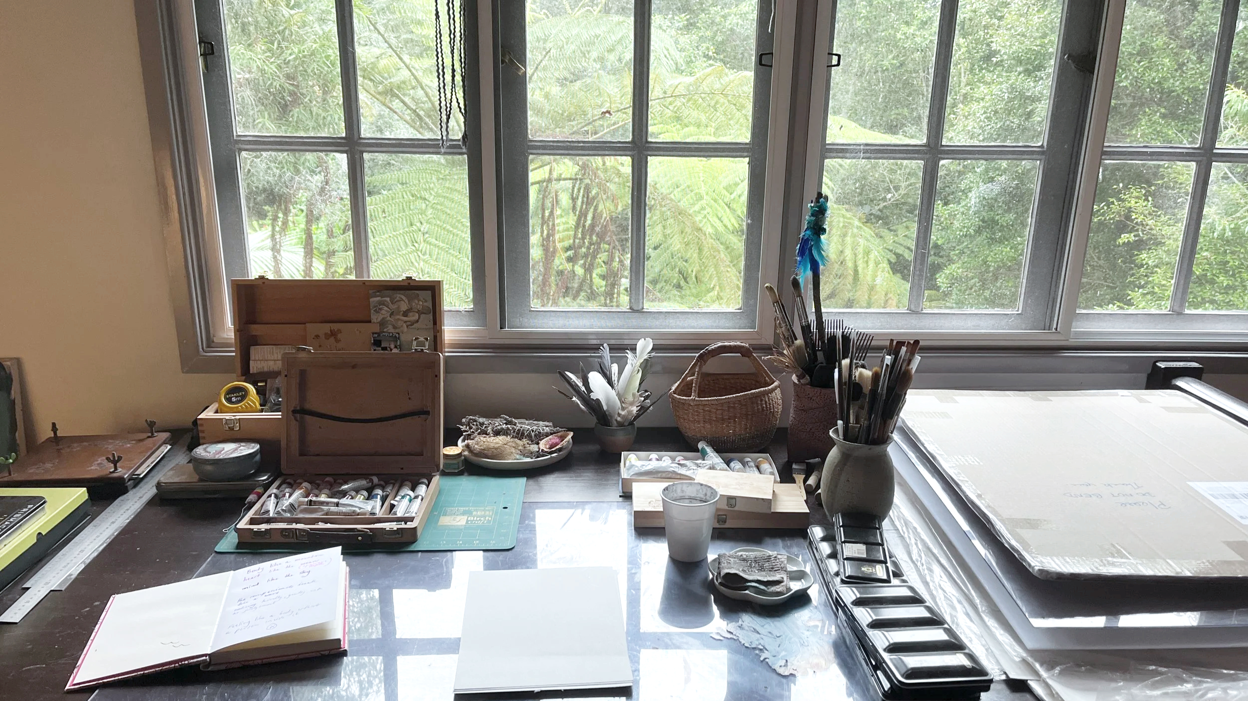 Art studio workspace with various art supplies, a notebook, and a window overlooking lush greenery outside.