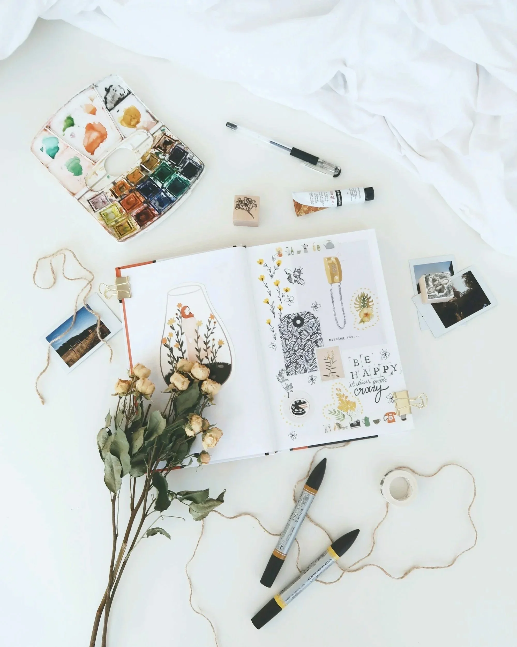Open notebook with colorful drawings and quotes, art supplies including watercolor paints, markers, and stickers, dried flowers, Polaroid photos, roll of tape, pens, and twine on a white surface, with crumpled white paper in the background.