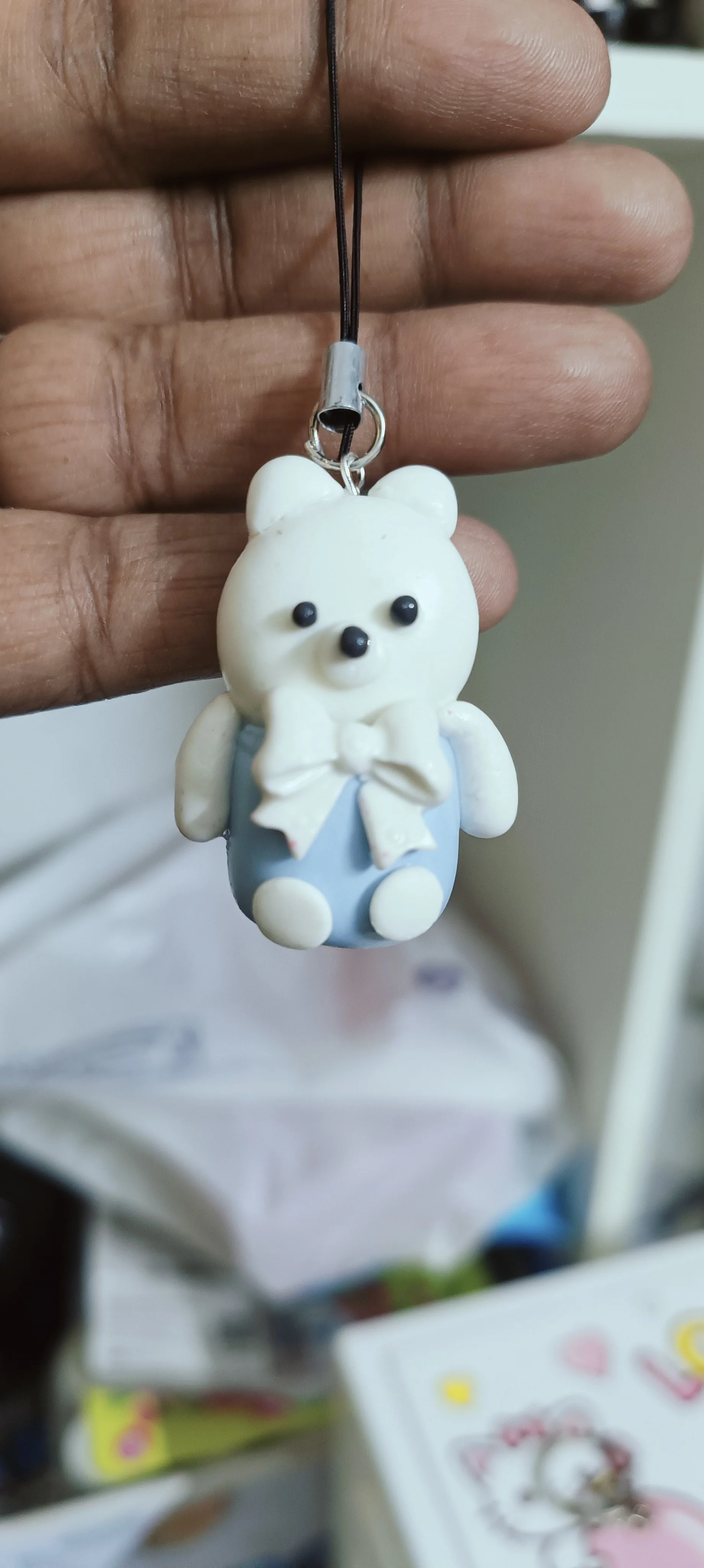 A small bear charm with a white face, black eyes and nose, wearing a blue outfit with a white bow, hanging from a black strap.