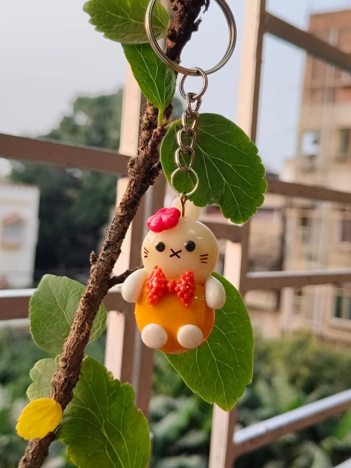 A keychain featuring a small figurine of Hello Kitty with a red bow, red bowtie, and white and yellow body, hanging from a plant stem with green leaves, against a blurred outdoor background.