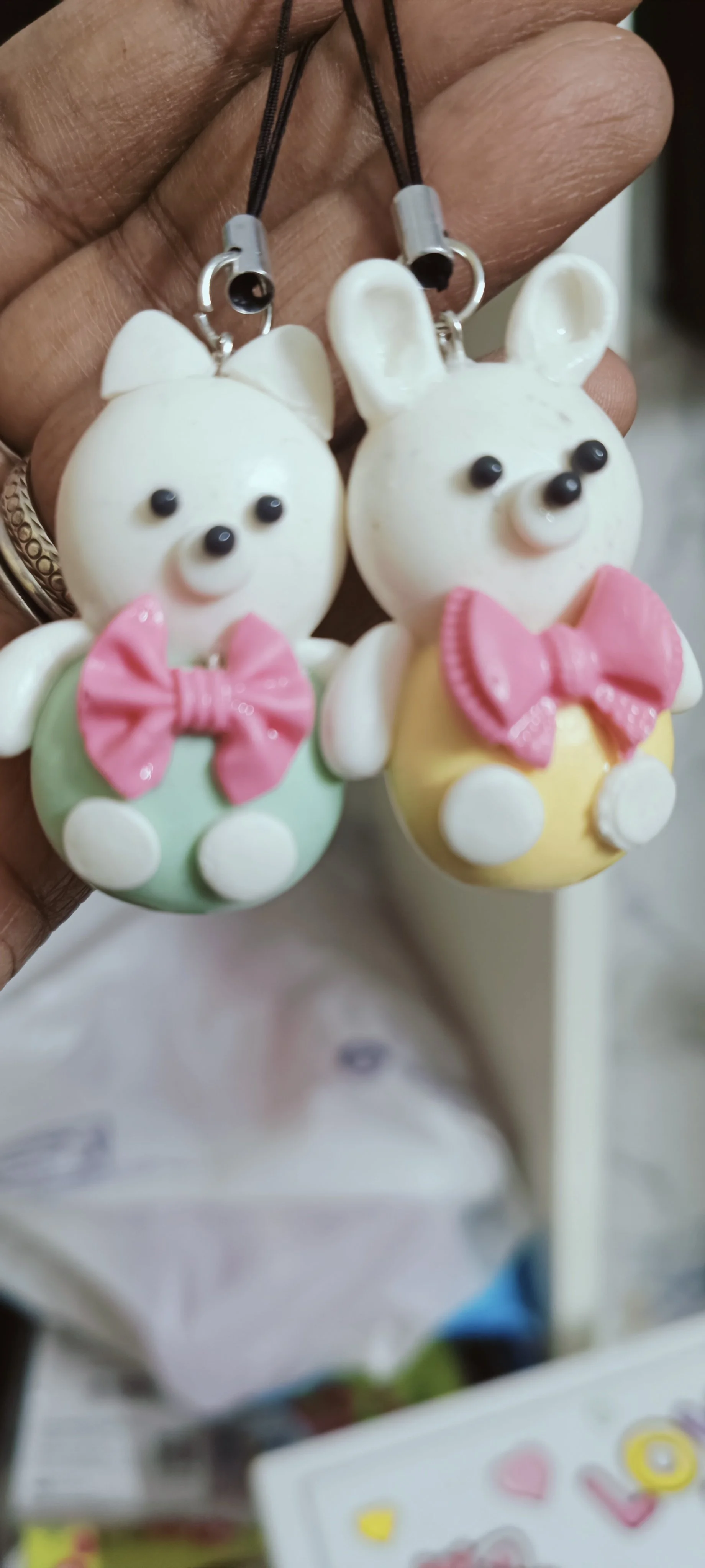 Colorful animal-shaped keychains or charms, resembling a bear and a rabbit, with bow ties, held up by a hand.