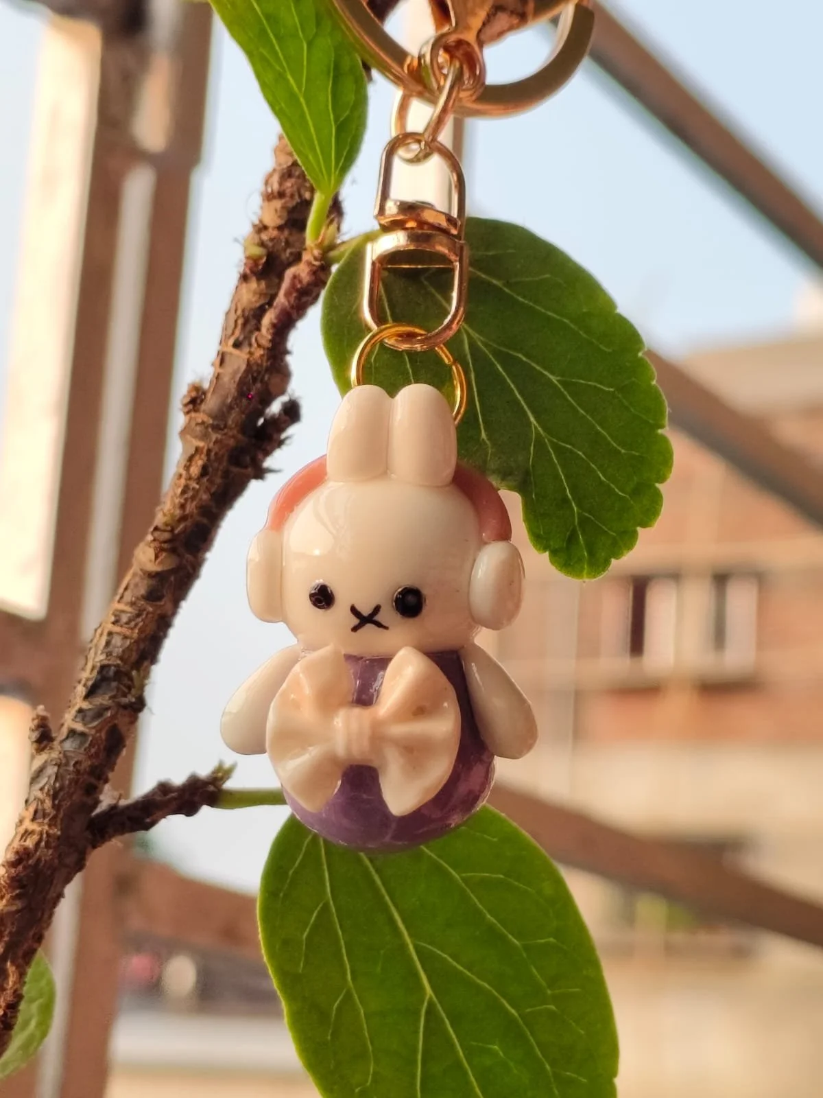 Close-up of a small plush keychain hanging from a leaf on a plant, with a house in the blurred background.