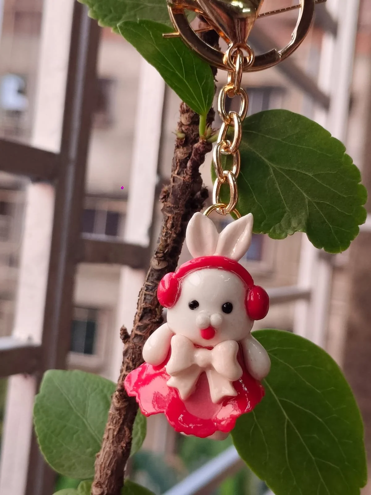 A keychain with a bunny figurine wearing red headphones, attached to a leafless branch with green leaves, in front of a building with multiple windows.