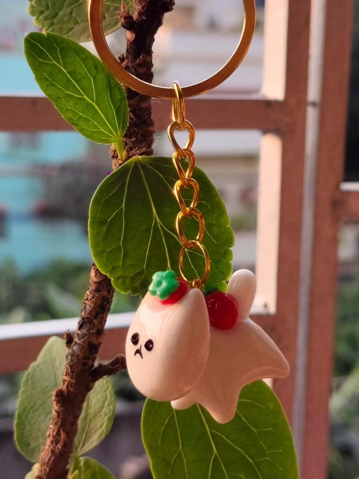 A keychain hanging from a leafy plant branch with a blurred background of buildings. The keychain has a gold chain and ring with a white, cute, smiling ghost-shaped figure adorned with tiny red and green decorations.