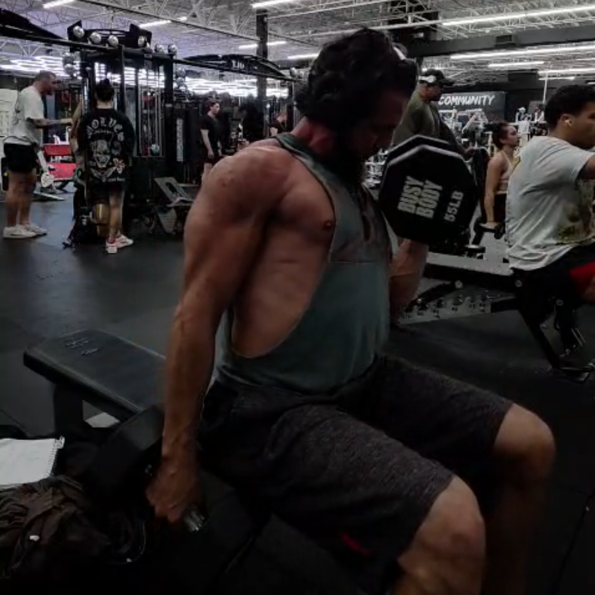 A muscular man in a gym, sitting on a workout bench, holding a dumbbell behind his back during strength training.