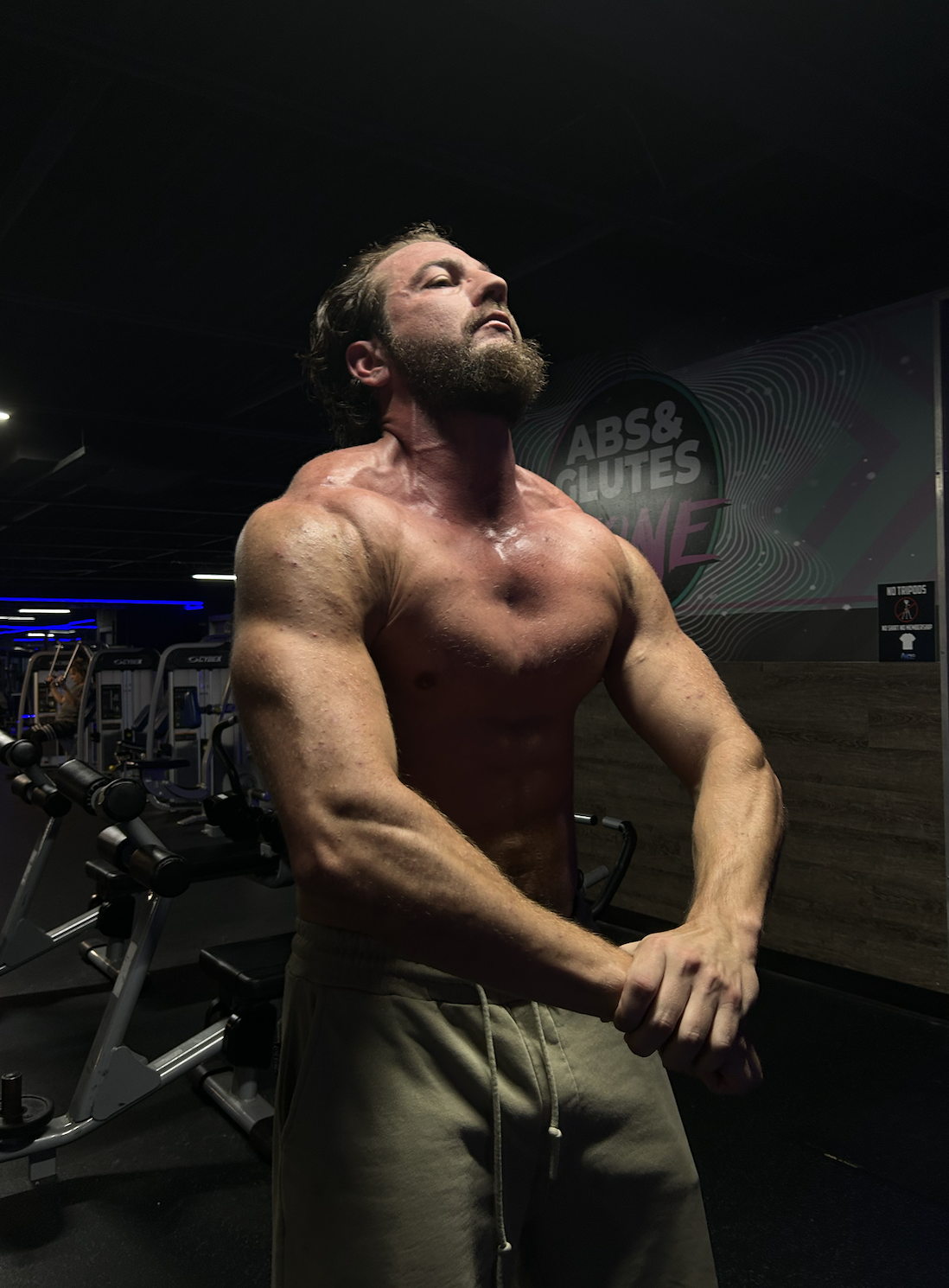 A muscular man with a beard flexing his biceps in a gym, with dark lighting and gym equipment in the background.