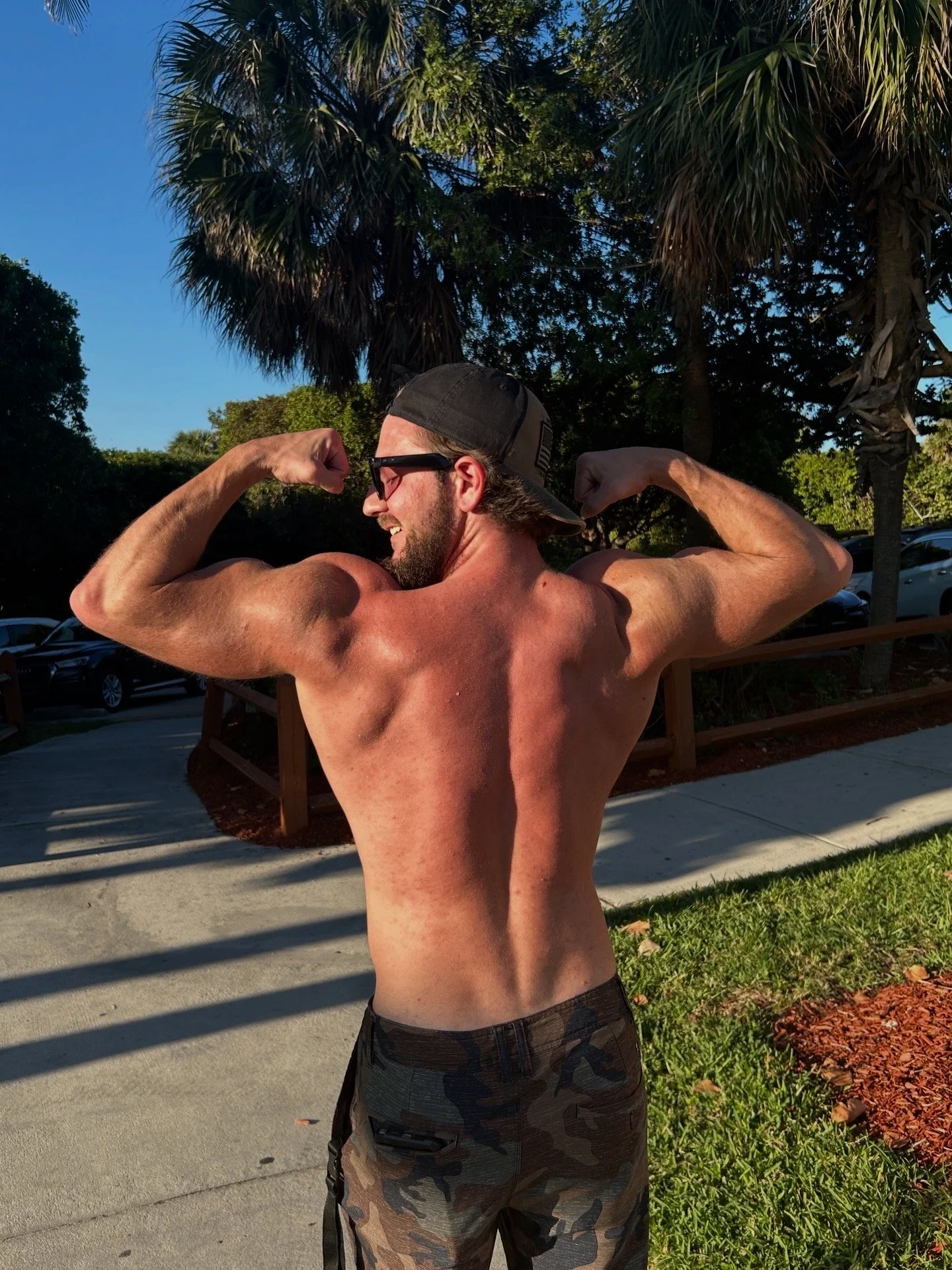 A shirtless man with long hair, dark sunglasses, and a backward cap is flexing his biceps outdoors on a sunny day, surrounded by palm trees, grass, a sidewalk, and parked cars.
