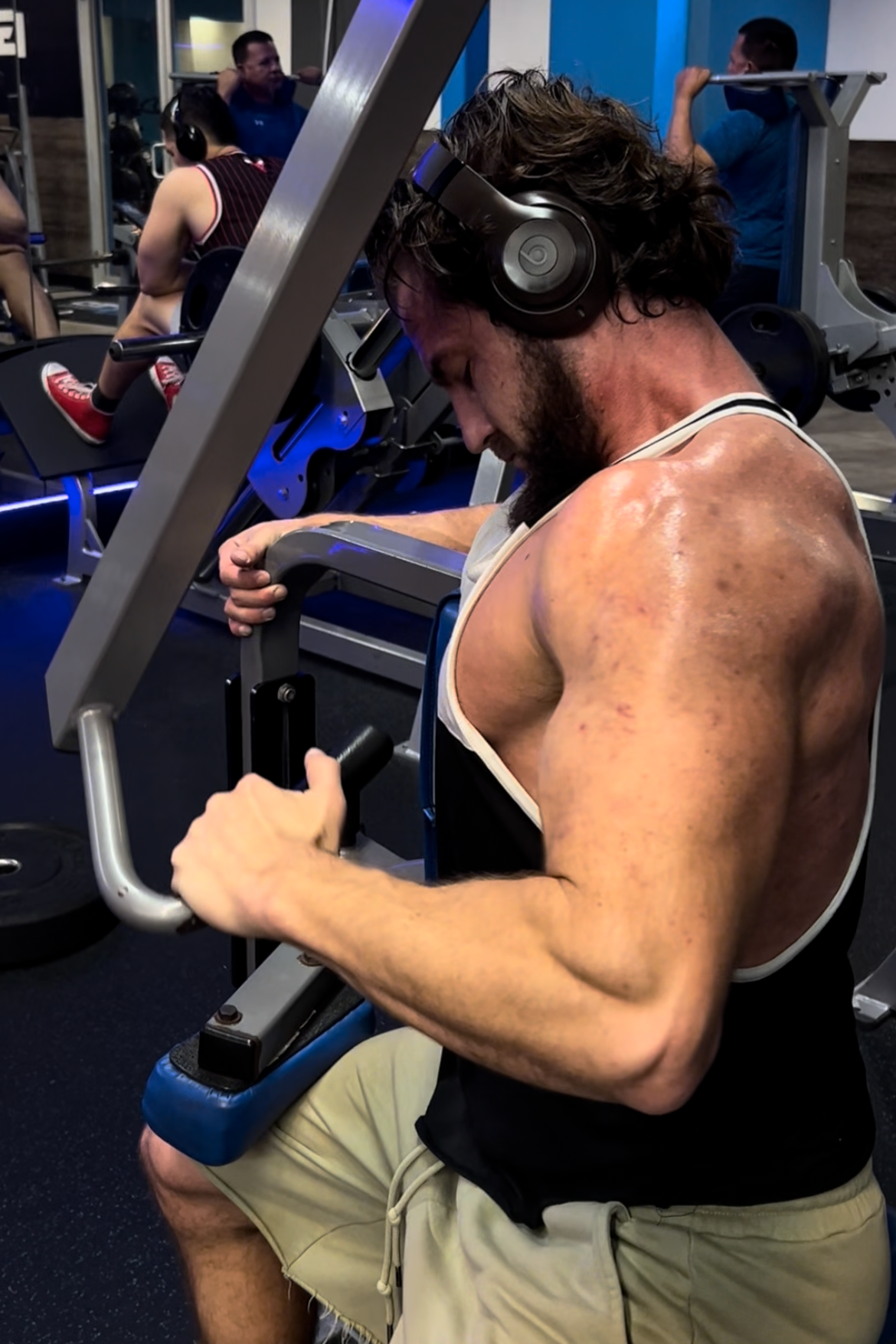 A man with a beard and headphones working out on a seated leg curl machine at the gym, with other people exercising in the background.
