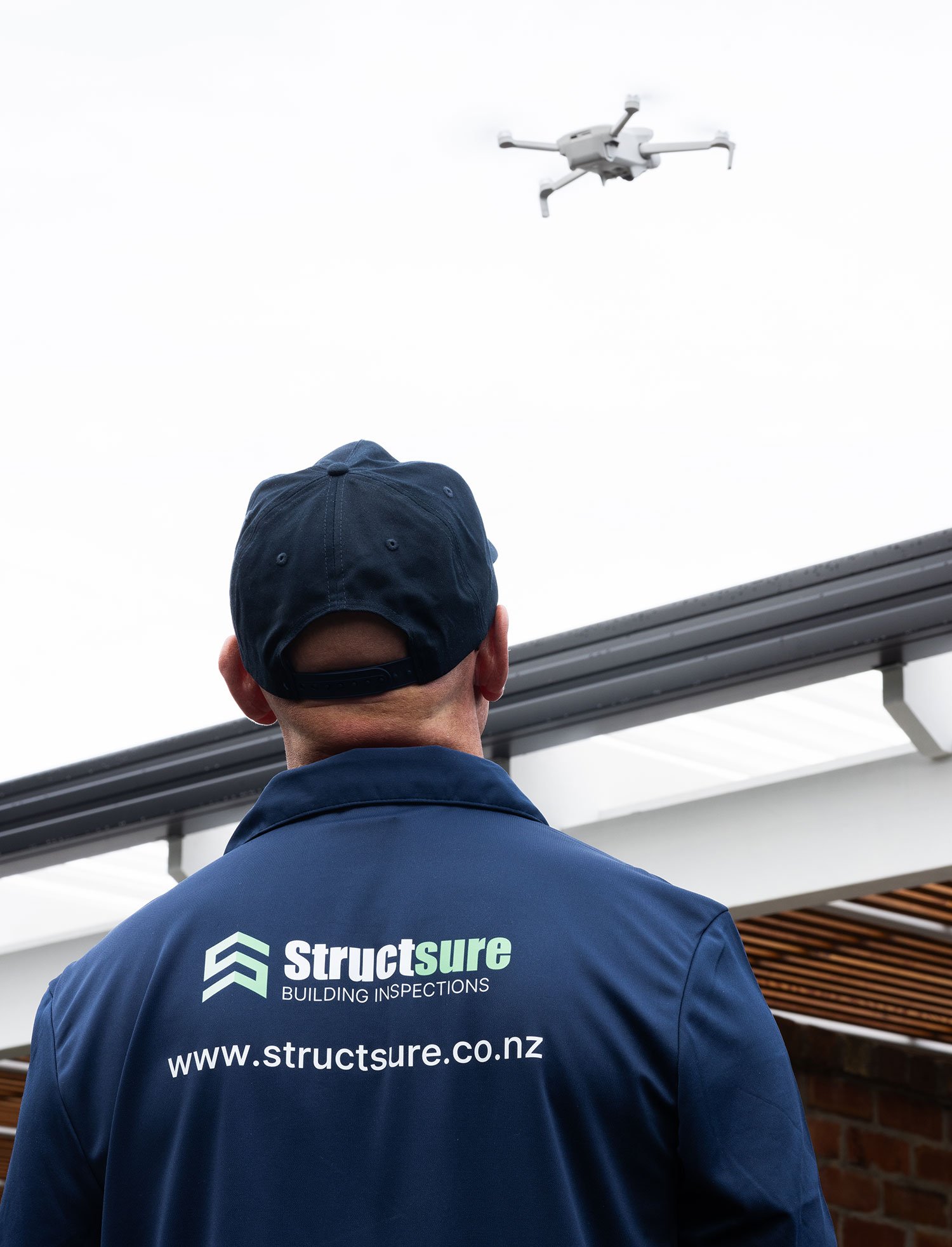 A person wearing a blue jacket and a navy baseball cap, standing outdoors and looking up at a drone flying in the cloudy sky. The jacket has a logo and the website 'www.structsure.co.nz' printed on the back.