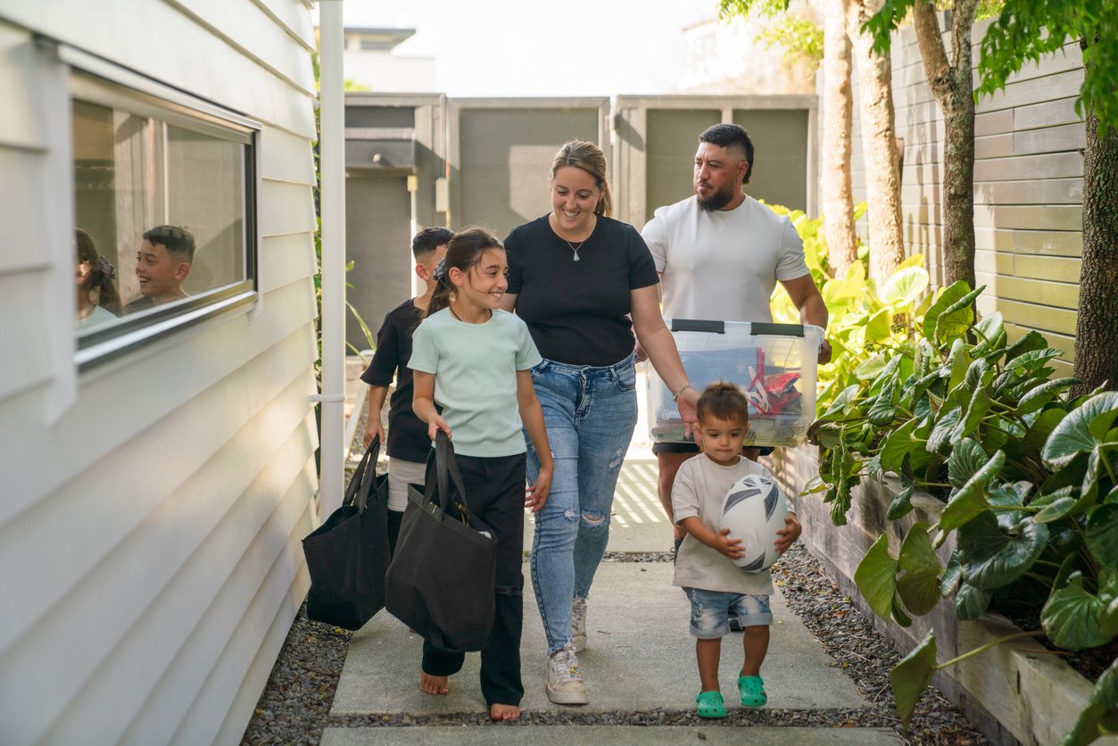 A family of six, including two children, walking outside in the backyard. They are carrying bags, a storage bin, and a ball, smiling and happy.