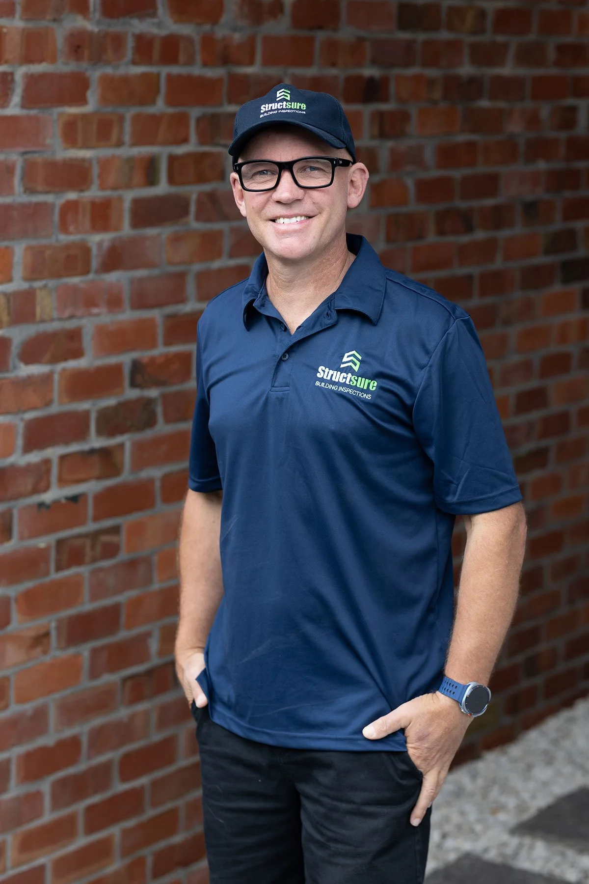 A man wearing glasses, a navy blue baseball cap, and a navy blue polo shirt with the 'Structsure' logo stands in front of a brick wall, smiling at the camera.